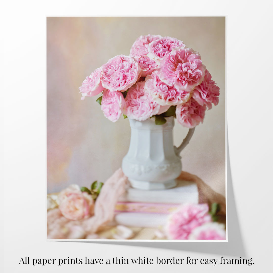 The Miranda Muse paper print features a white ceramic pitcher filled with pink peonies atop a stack of books, surrounded by blooms. It includes a thin white border, as do all paper prints, for easy framing.