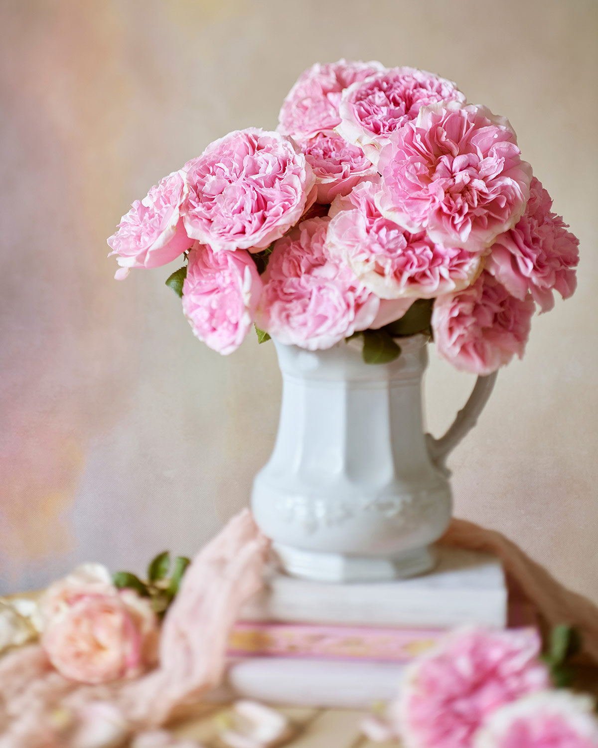 A white ceramic pitcher filled with Miranda roses sits atop a stack of books. The softly blurred pastel background and light pink cloth draped around the base enhance the romantic, vintage aesthetic.