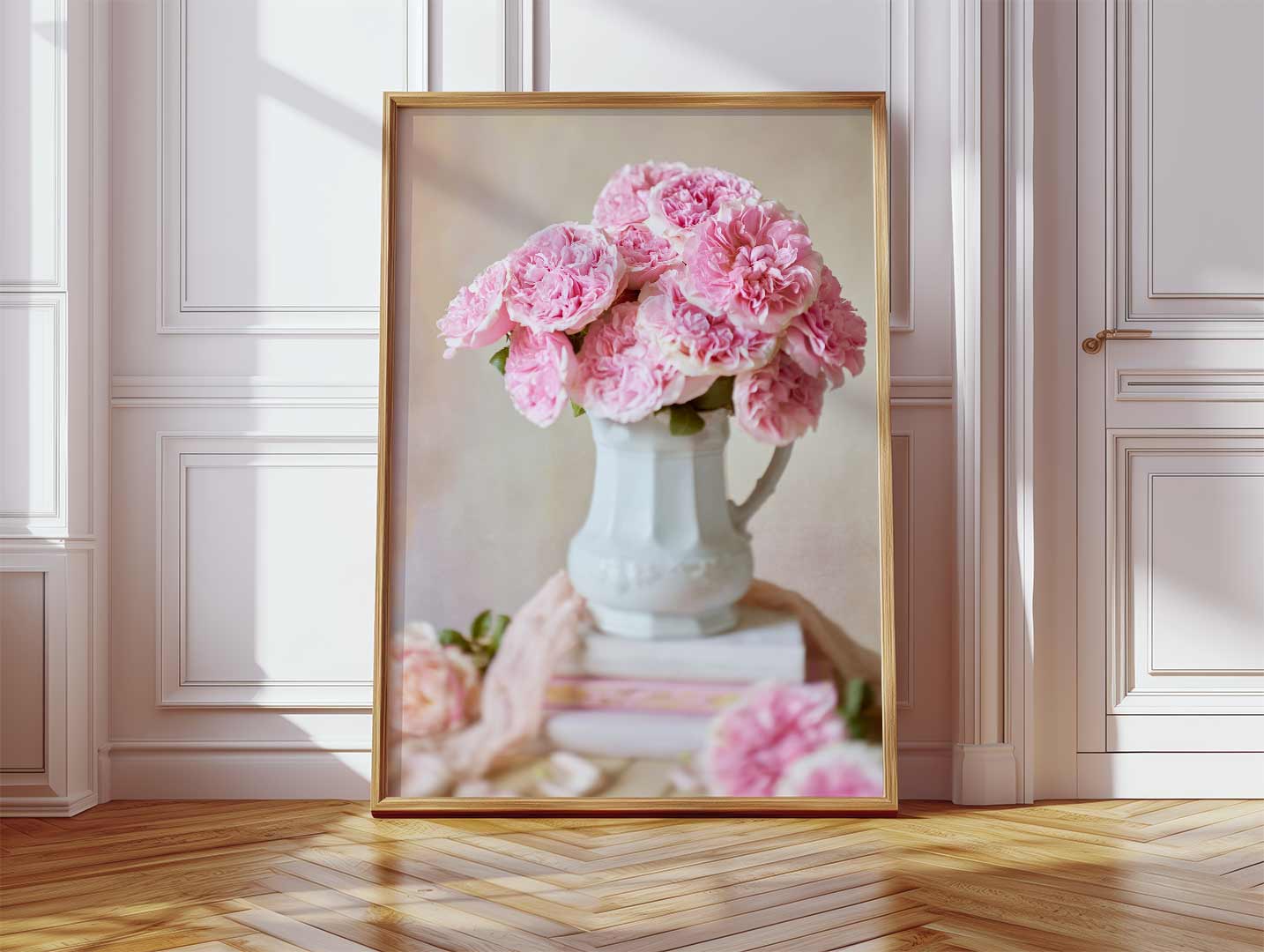 A large framed canvas print, Miranda Muse, showcasing pink Miranda David Austin roses in a white vase leans against a wooden floor. The softly lit room features white paneled walls with light streaking across the canvas from the window.