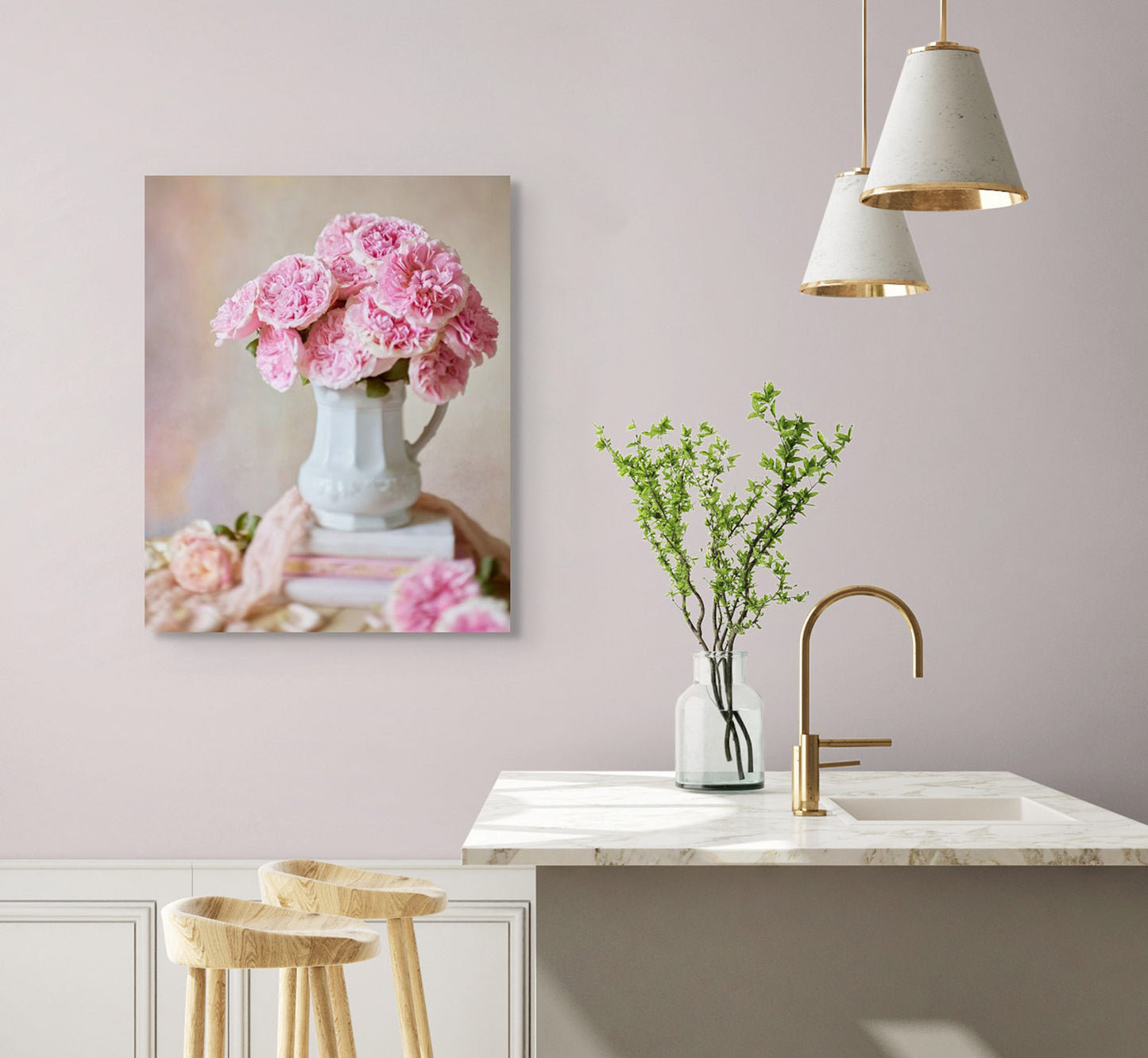 A minimalist kitchen with light pink walls features a marble countertop, a vase with green branches, and wooden stools. Canvas wall art of Miranda Muse roses adorns the wall. Two pendant lights hang gracefully above the counter, enhancing the serene ambiance.