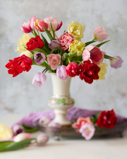 Spring Symphony, a white vase filled with vibrant red, pink, yellow, and purple tulips spilling over the rim, sits on a table. Scattered tulips and a purple cloth beside it create an elegant setting against a soft-focus gray backdrop.