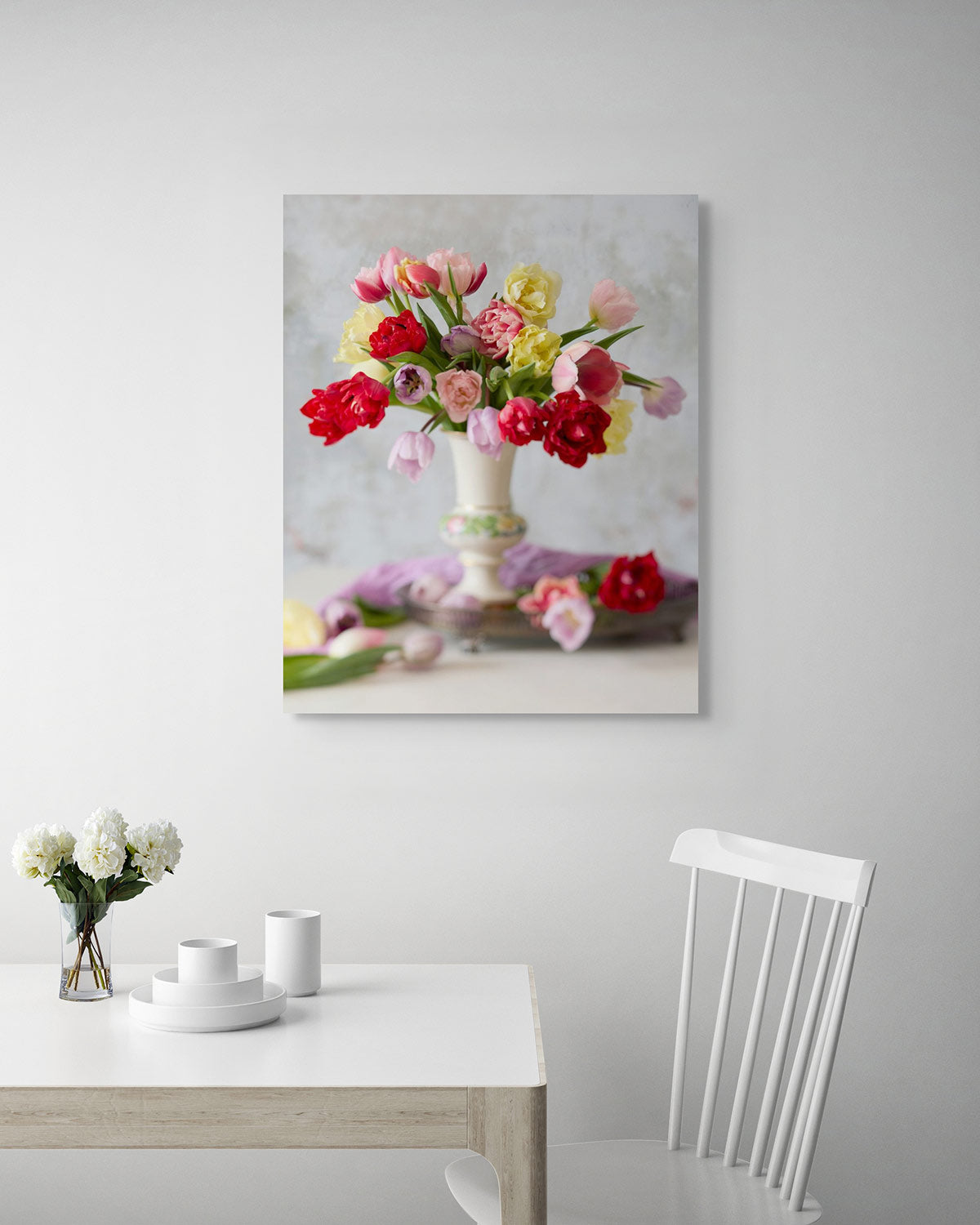 A white dining area features a wooden table, a white chair, and a vase of white flowers. The space is enlivened by Spring Symphony, a canvas art piece exhibiting vibrant tulip wall art, showcasing colorful tulips in a vase on the wall.