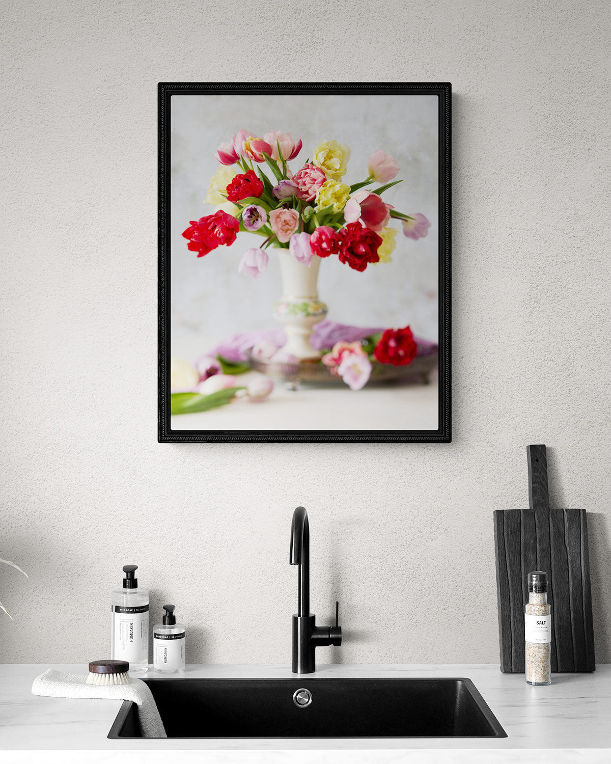 A modern kitchen interior displays a matte black sink faucet and soap dispensers on a white countertop. Above the sink, Spring Symphony wall art showcases vibrant tulips in red, pink, and yellow in a decorative vase, with wooden boards leaning against the wall.