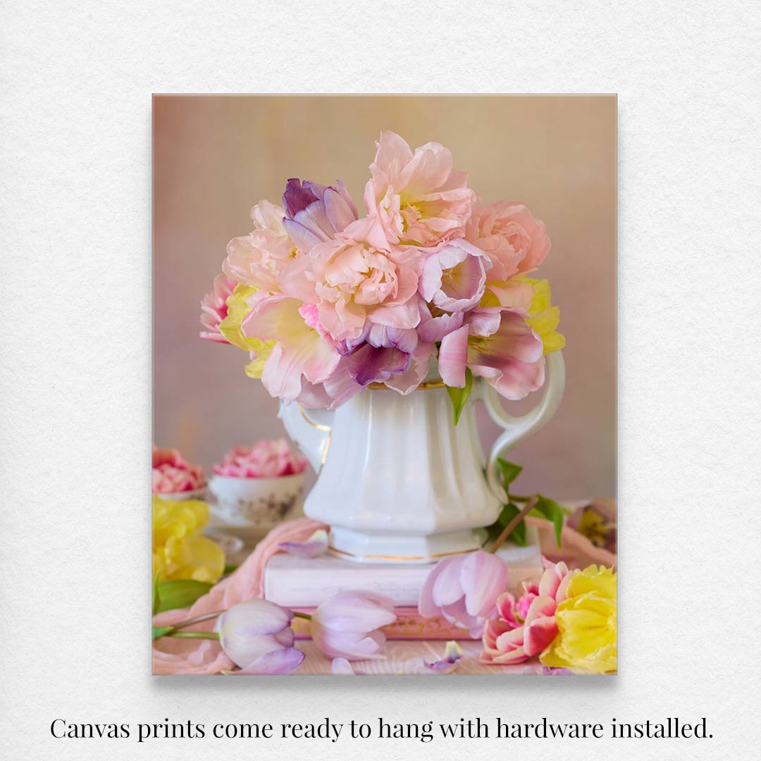 The Teapot of Blooms fine art print displays a white vase of pink, yellow, and purple peonies alongside spring tulips against a soft backdrop, with extra flowers on top of a few books. The text states that canvas prints are ready to hang with hardware installed.