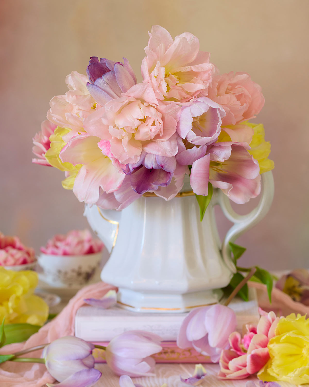 A white vase holds a bouquet of spring tulips in pastel pink, purple, and yellow. This scene captures the essence of the Teapot of Blooms canvas wall art, set on a table with scattered flowers and a soft-focus background for a delicate romantic composition.