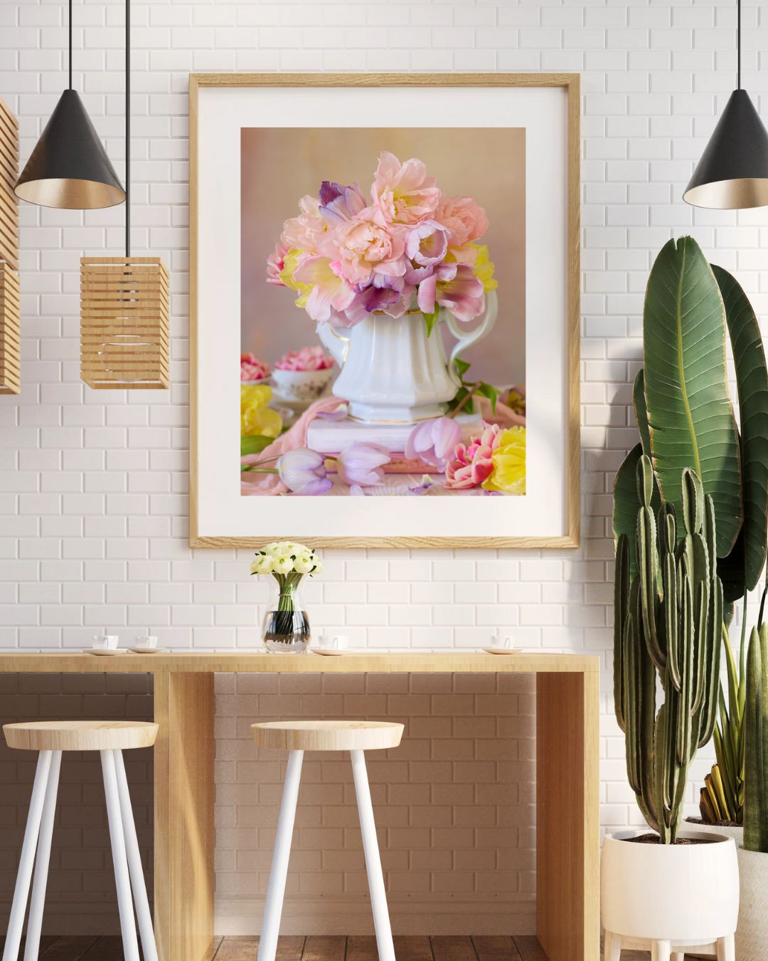 A modern dining area features a light wood table with two stools beneath it. A framed print, Teapot of Blooms, hangs on a white brick wall. The space is accented by hanging lamps and a tall potted plant.