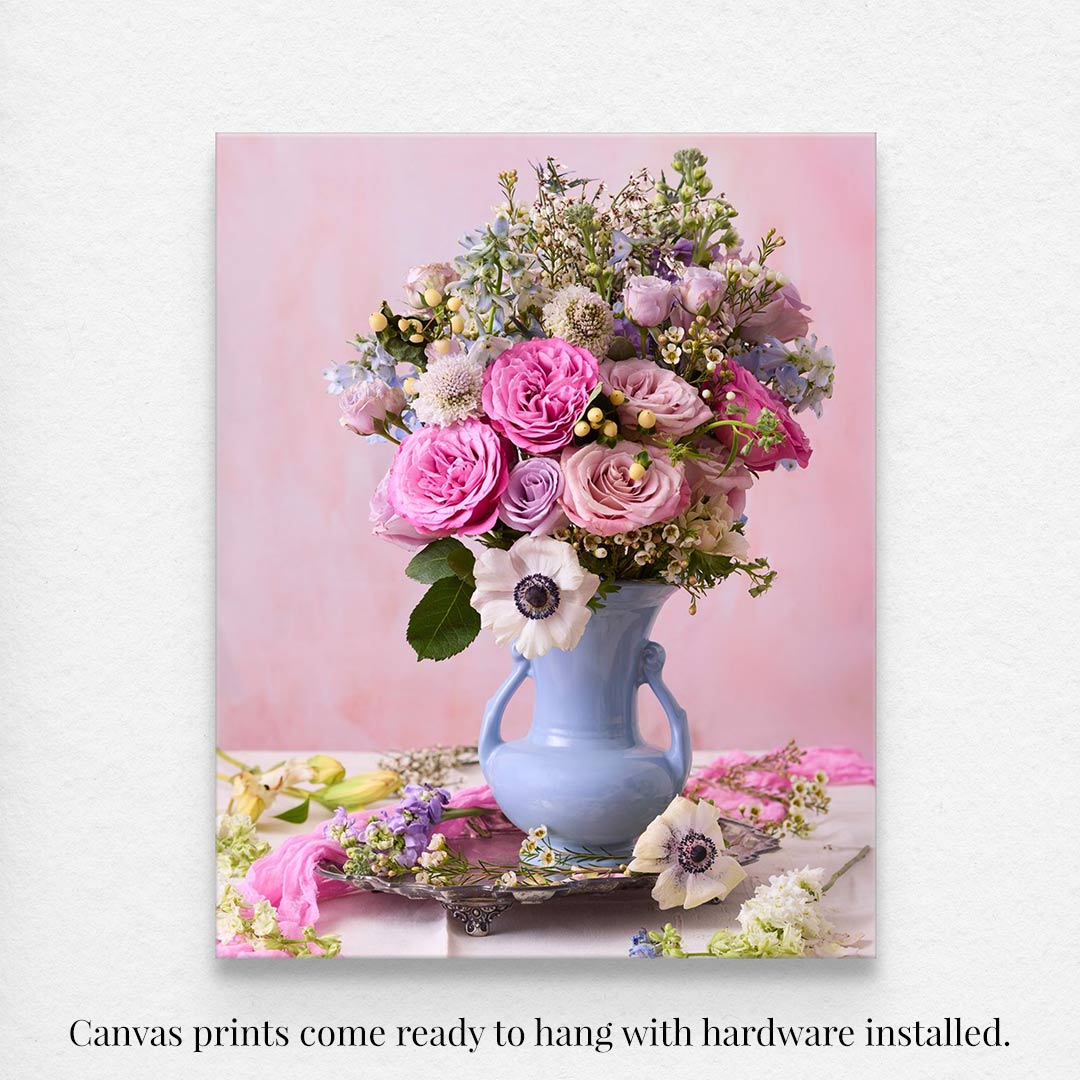 The Pink and Blue Serenade canvas art showcases a bouquet of pastel roses, purple flowers, and greenery in a light blue vase on a soft pink background. It comes ready to hang with hardware installed.