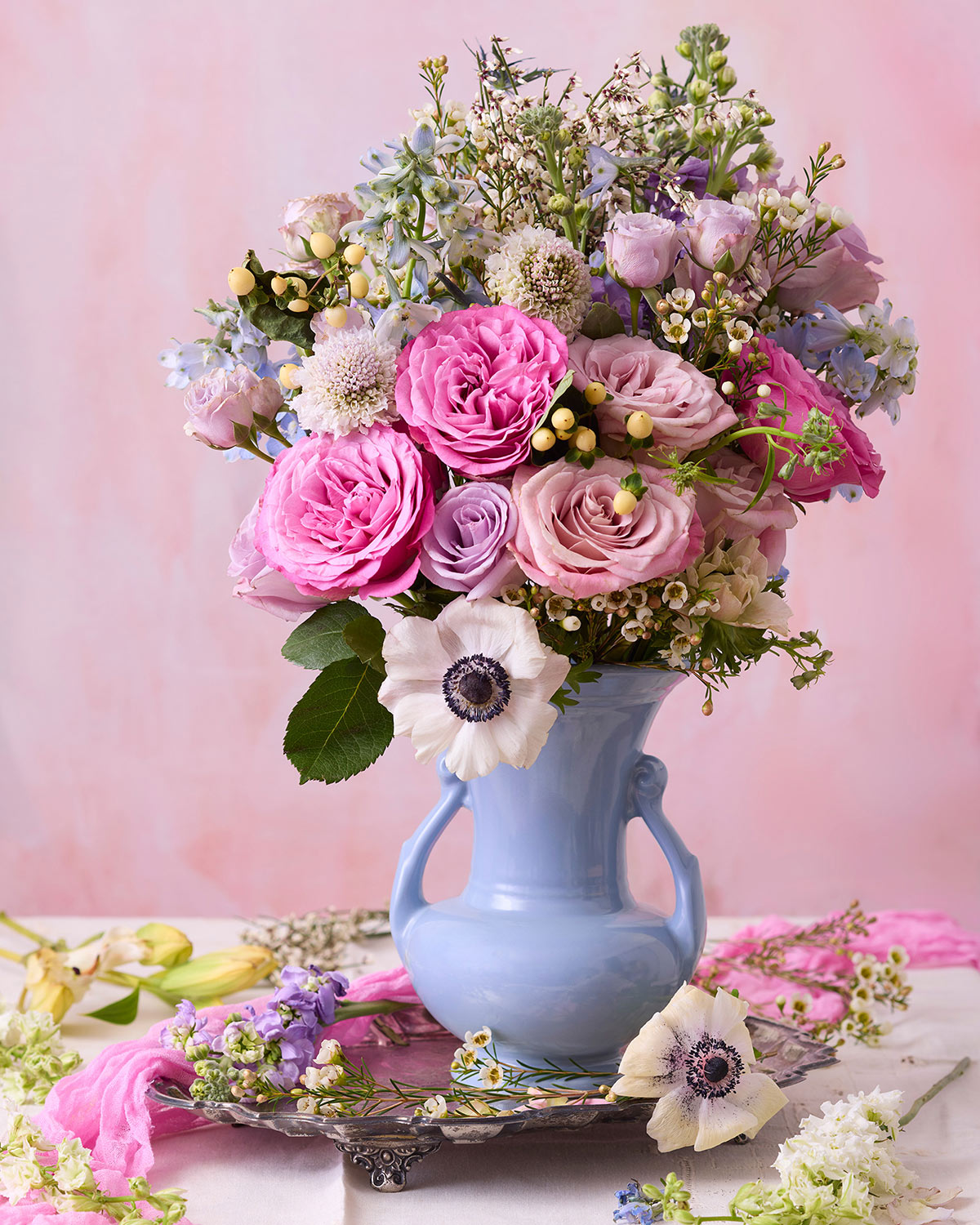 The Pink and Blue Serenade, features pink pastel roses, white anemones, and blue and purple flowers in a light blue vase. Set on a decorative tray against a soft pink backdrop with loose flowers scattered around.