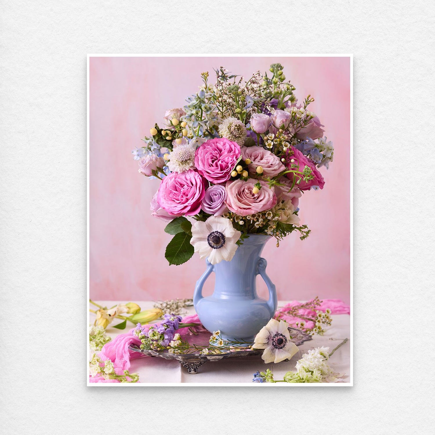 The pastel floral arrangement, Pink and Blue Serenade, features pink roses, purple flowers, and white anemones in a light blue vase. Set against a soft pink background with scattered petals on the table. Paper prints have a thin white border for easy framing.