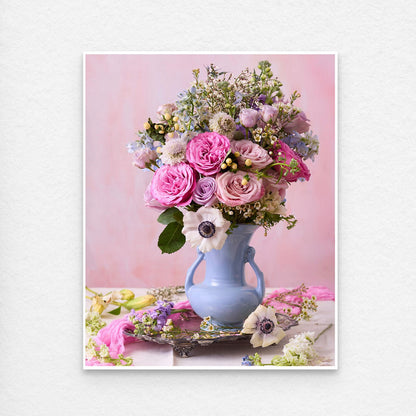 The pastel floral arrangement, Pink and Blue Serenade, features pink roses, purple flowers, and white anemones in a light blue vase. Set against a soft pink background with scattered petals on the table. Paper prints have a thin white border for easy framing.