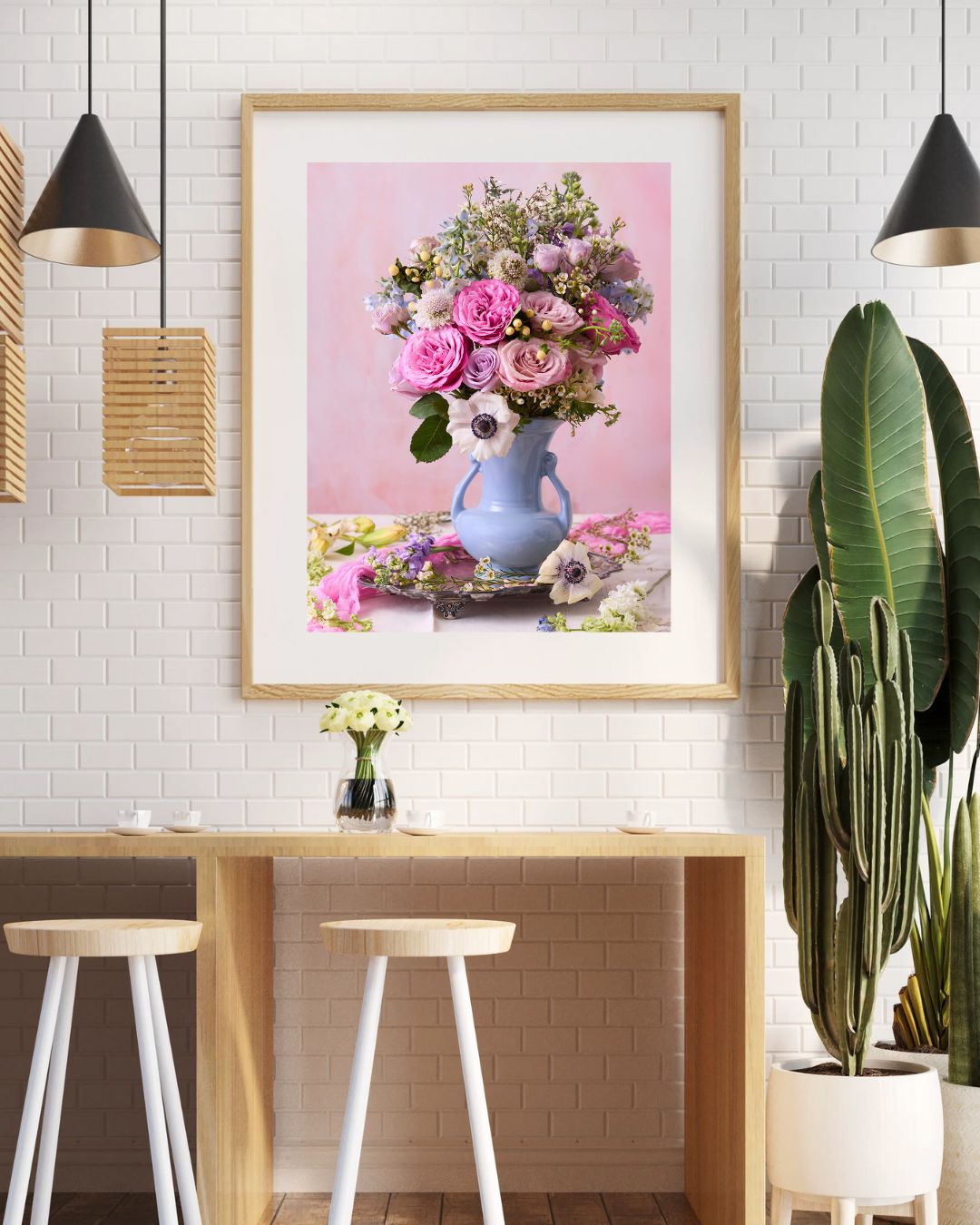 The cafés cozy interior features a wooden counter with two white stools. Above, the Pink and Blue Serenade framed print of a floral bouquet in a blue vase adds sophistication to the textured white wall, complemented by a tall cactus on the right.