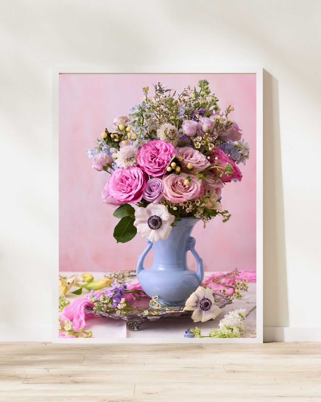 The Pink and Blue Serenade framed canvas showcases a pastel floral arrangement in a light blue vase with pink roses, white anemones, and lush greenery against a soft pink backdrop. The canvas is leaning on a white wall.