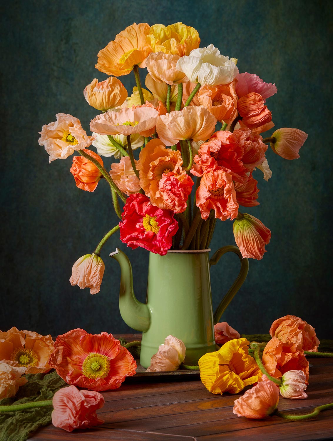 The Poppy Sisters Portraits - 3 Piece features a vibrant Italian pastel bouquet of orange, yellow, and pink poppies elegantly arranged in a green teapot vase with some petals tastefully scattered on the wooden surface below, set against a textured dark background.