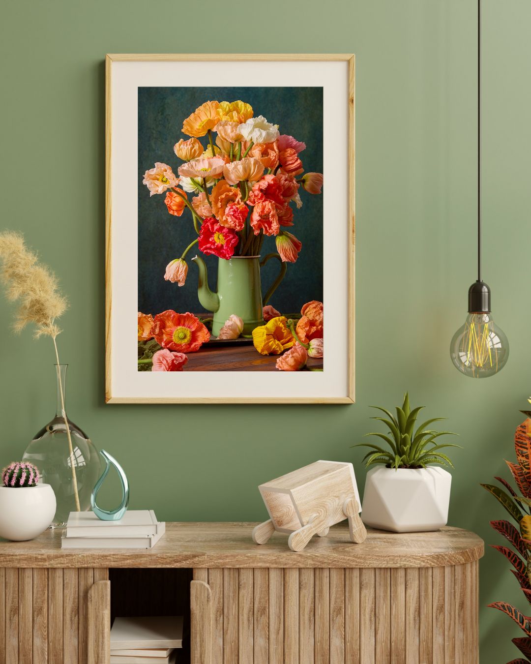 A wooden console with books, decor, and plants stands against a green wall. Above hangs the vibrant Poppy Passion print framed with a white mat.