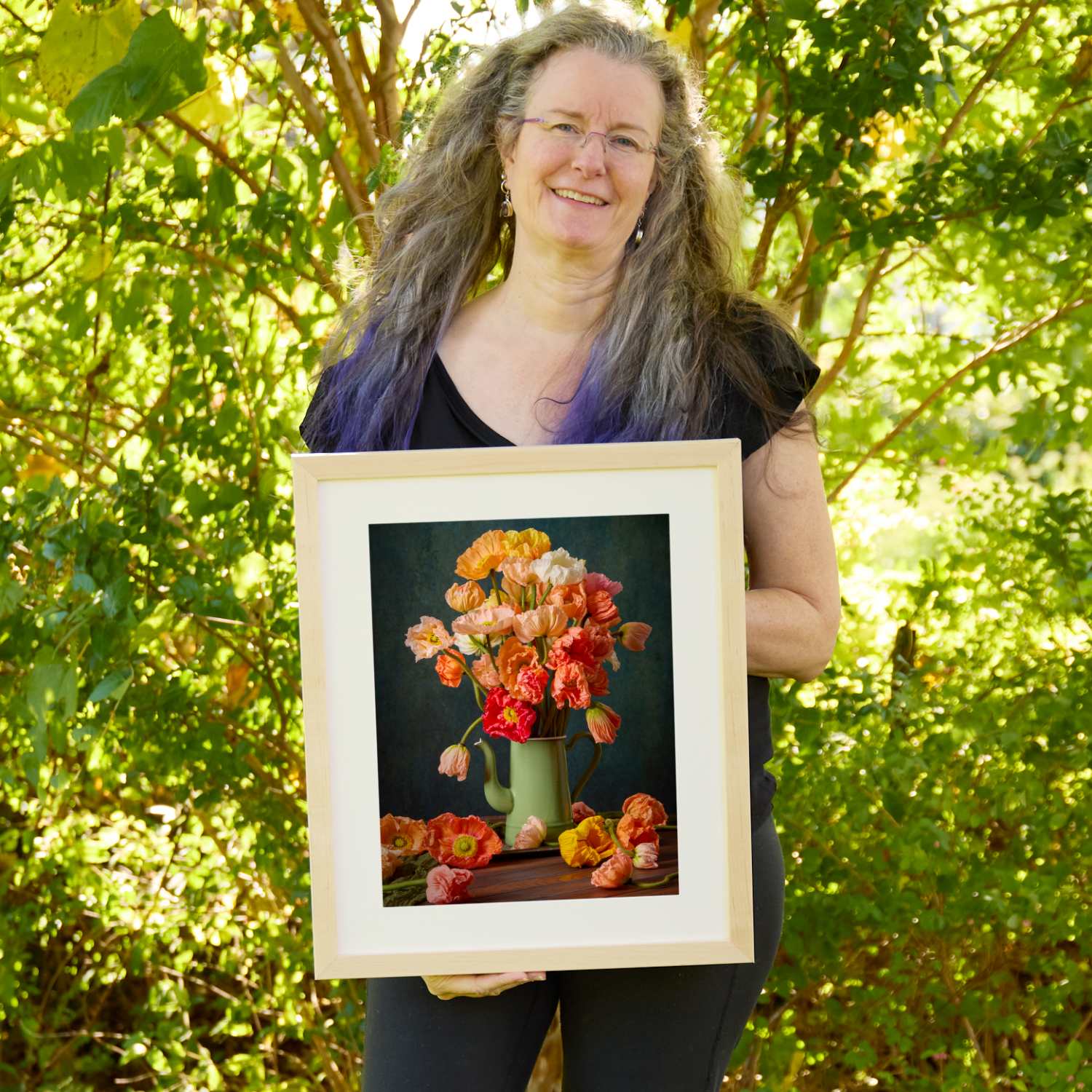 Christina Peters holding Poppy Passion Wall Art Print, a still life of orange, coral, red and cream Iceland poppies in a sage green enamel coffee pot, with fallen poppies on the table against a dark teal backdrop, framed in natural wood with a white mat. She is standing outdoors in front of sunlit green trees and leafy shrubs.