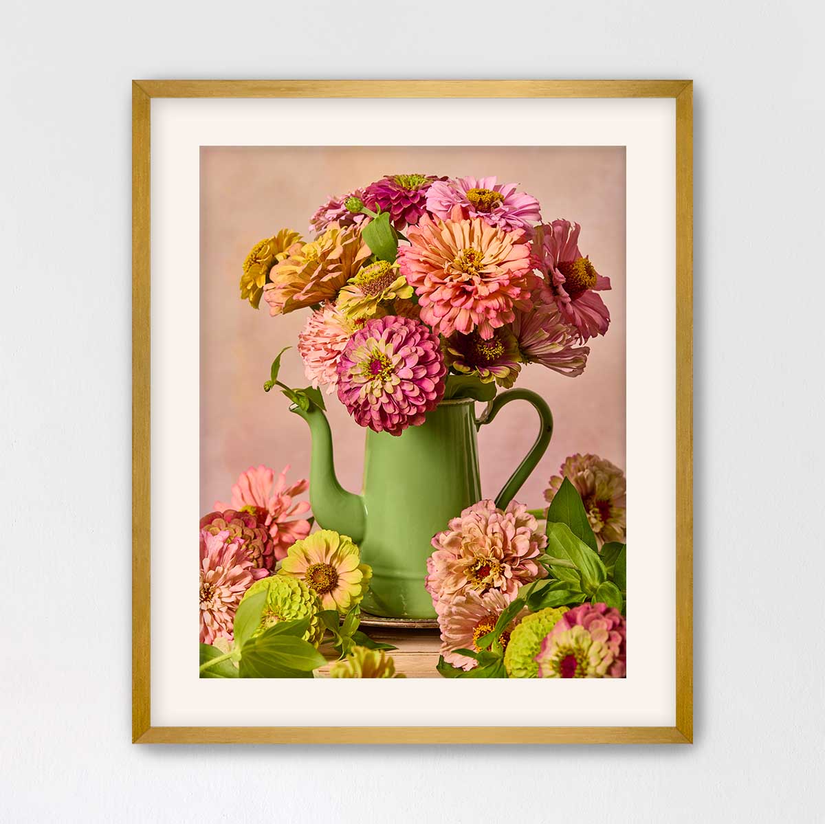 A framed image of a green teapot filled with a vibrant bouquet of pink and yellow flowers, placed on a wooden surface.