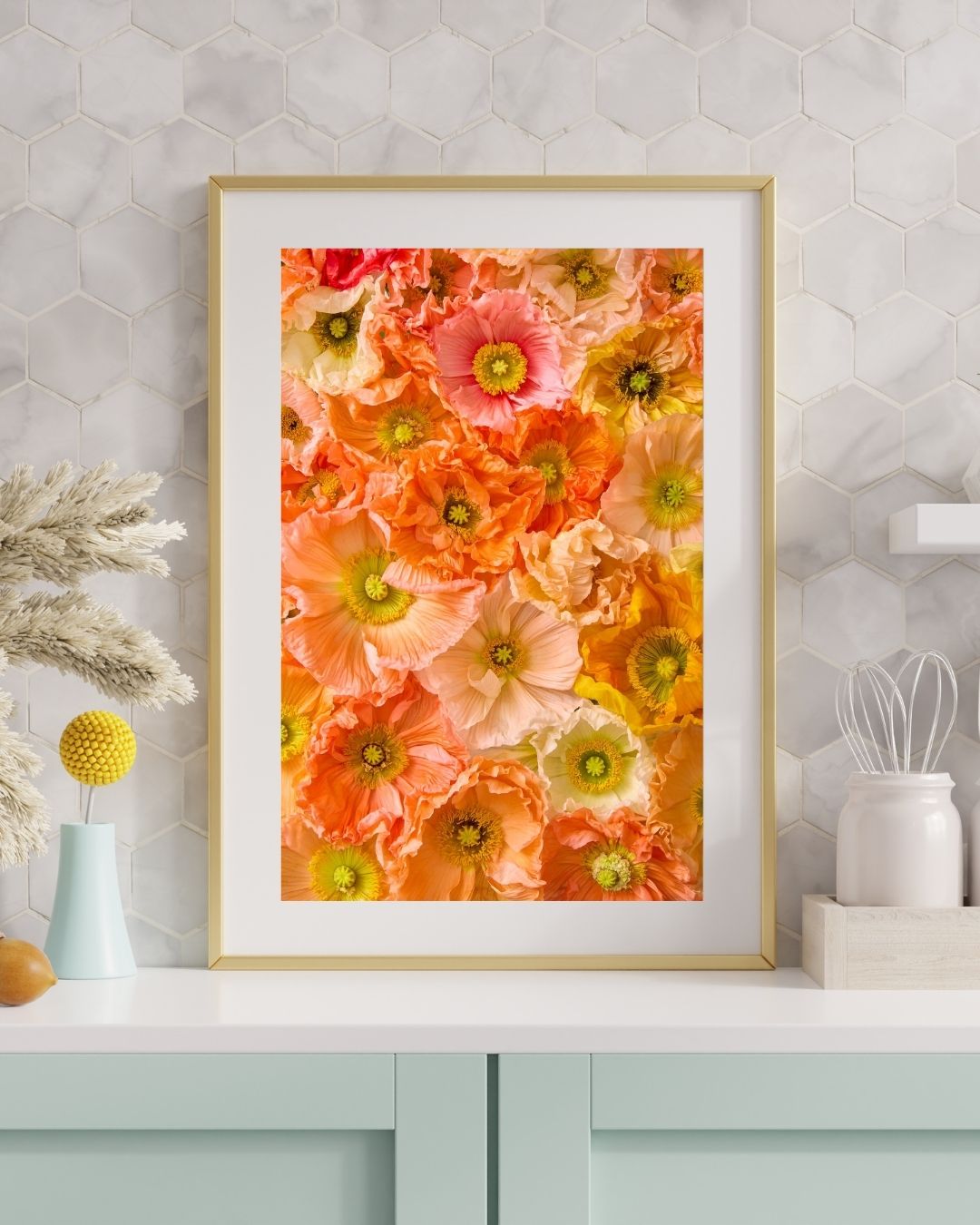 The Petals of Light framed artwork, showcasing a vibrant floral pattern of pink, orange, and yellow flowers like Italian pastel poppies, is displayed on a light-colored hexagonal-tiled wall. Nearby sits a vase with pampas grass, a yellow pom-pom flower, and white ceramic containers.