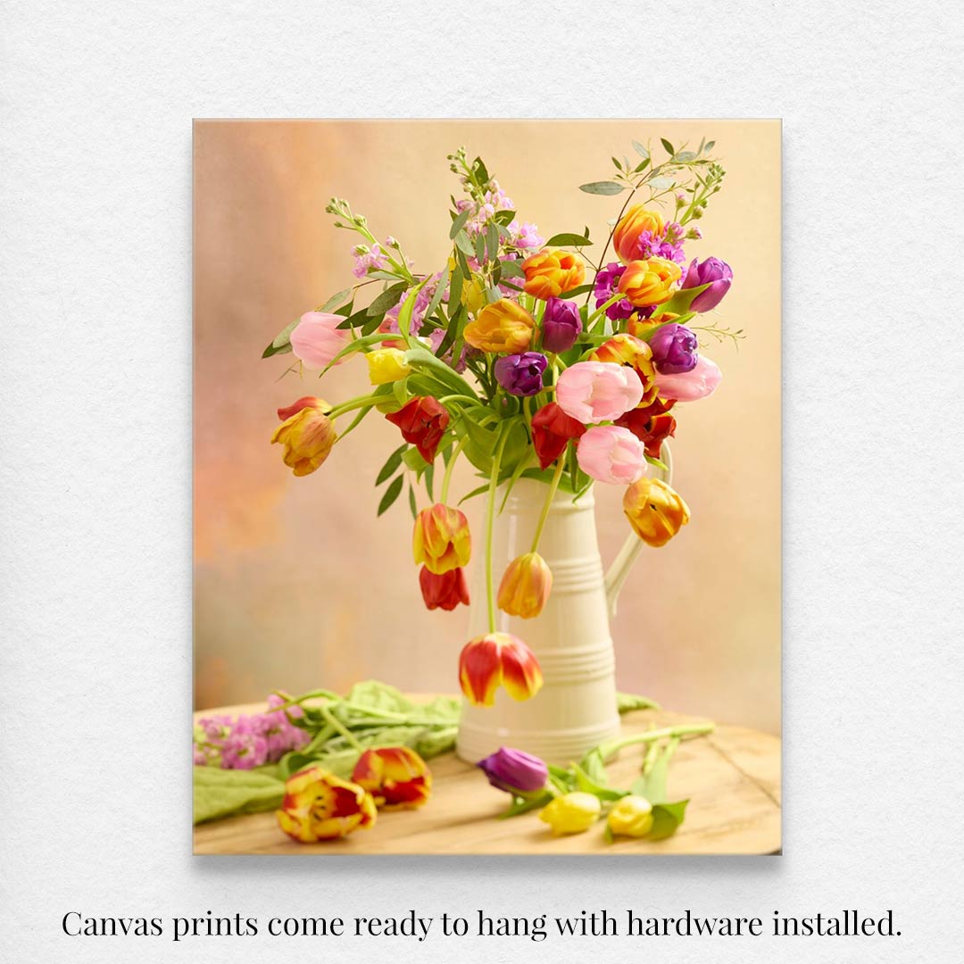 Tulip Cascade is a canvas wall art depicting a spring bouquet in a white jug, with vivid blooms spilling over the rim onto a light wooden surface. The background offers a soft pastel gradient, enhancing its casual charm. Text explaining the canvas art comes ready to hang.