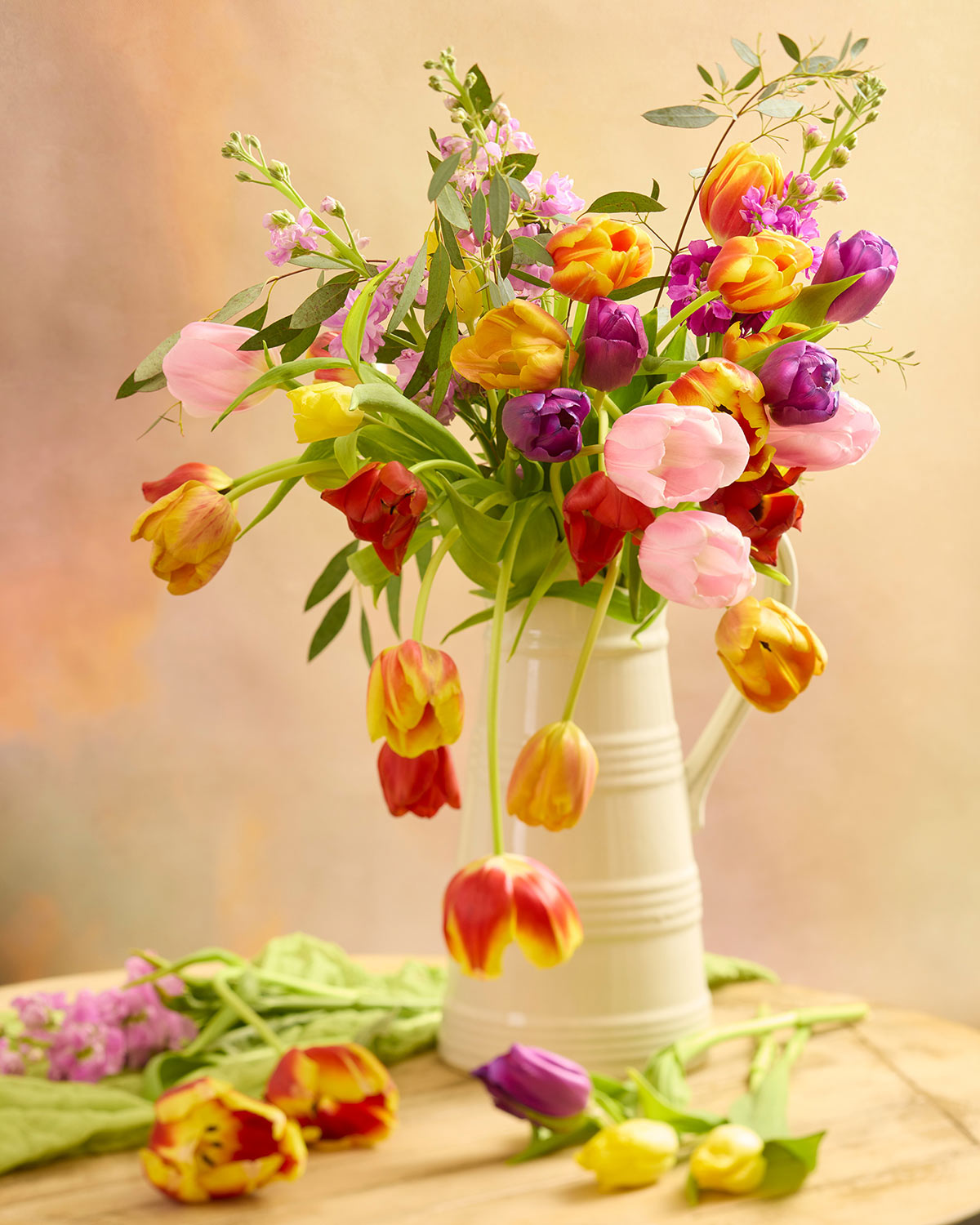 The Tulip Cascade, featuring vibrant pink, red, yellow, and purple tulips in a cream ceramic pitcher, is artfully arranged with loose tulips on a wooden surface against a soft pink and orange backdrop.