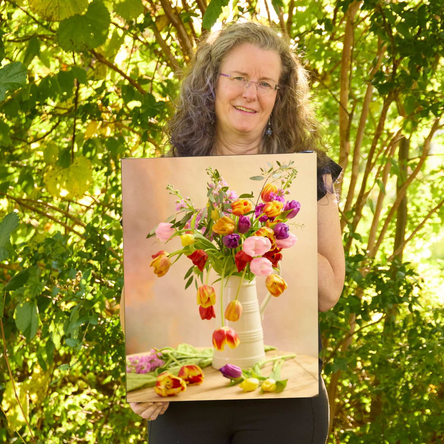 Christina Peters holding Tulip Cascade Wall Art Canvas, a still life of red, yellow, purple and pink tulips spilling from a cream pitcher, with loose tulips on the table against a warm peach backdrop. She is standing outdoors in front of sunlit green trees and leafy shrubs.