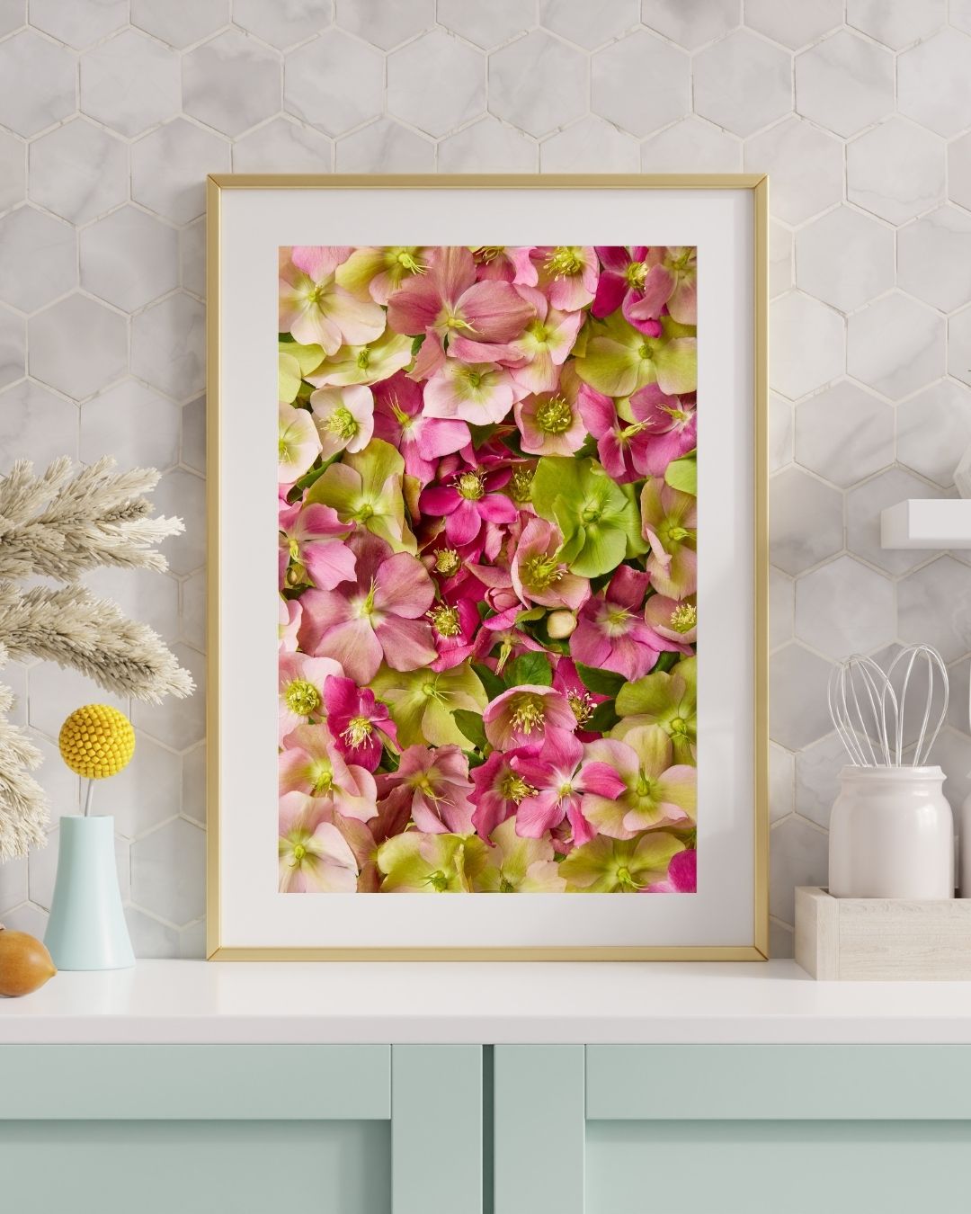 A matted and framed print of Hellebore Bed - Winter Roses graces a wall with hexagonal tiles. Below, a countertop displays a small vase of dried grasses, a yellow flower decoration, and a white kitchen utensil holder.