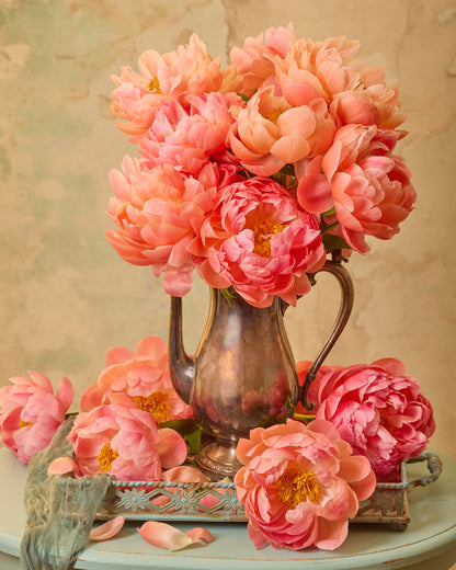 A vintage silver pitcher cradles a bouquet of vibrant Coral Charm peonies on a round table. Several loose flowers and petals are scattered around the base, with a soft green cloth partially draped on the surface. The backdrop features textured canvas wall art in soothing beige tones.