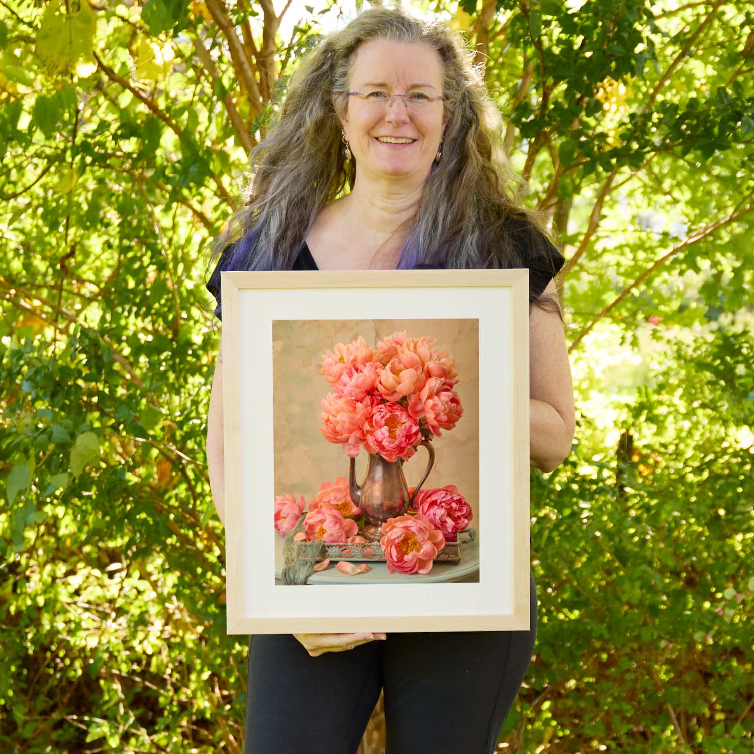 Christina Peters holding Coral Radiance Peony Wall Art Print, a still life of coral peonies in an antique copper pitcher on a vintage tray, with loose blooms at its base against a warm neutral backdrop, framed in natural wood with a white mat. She is standing outdoors in front of sunlit green trees and leafy shrubs.