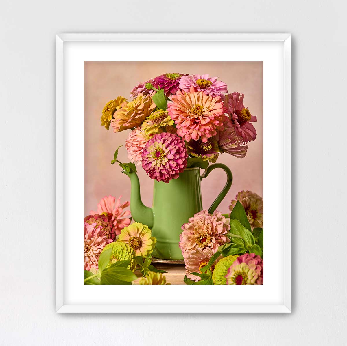 A framed image of a green teapot filled with a vibrant bouquet of pink and yellow flowers, placed on a white surface.