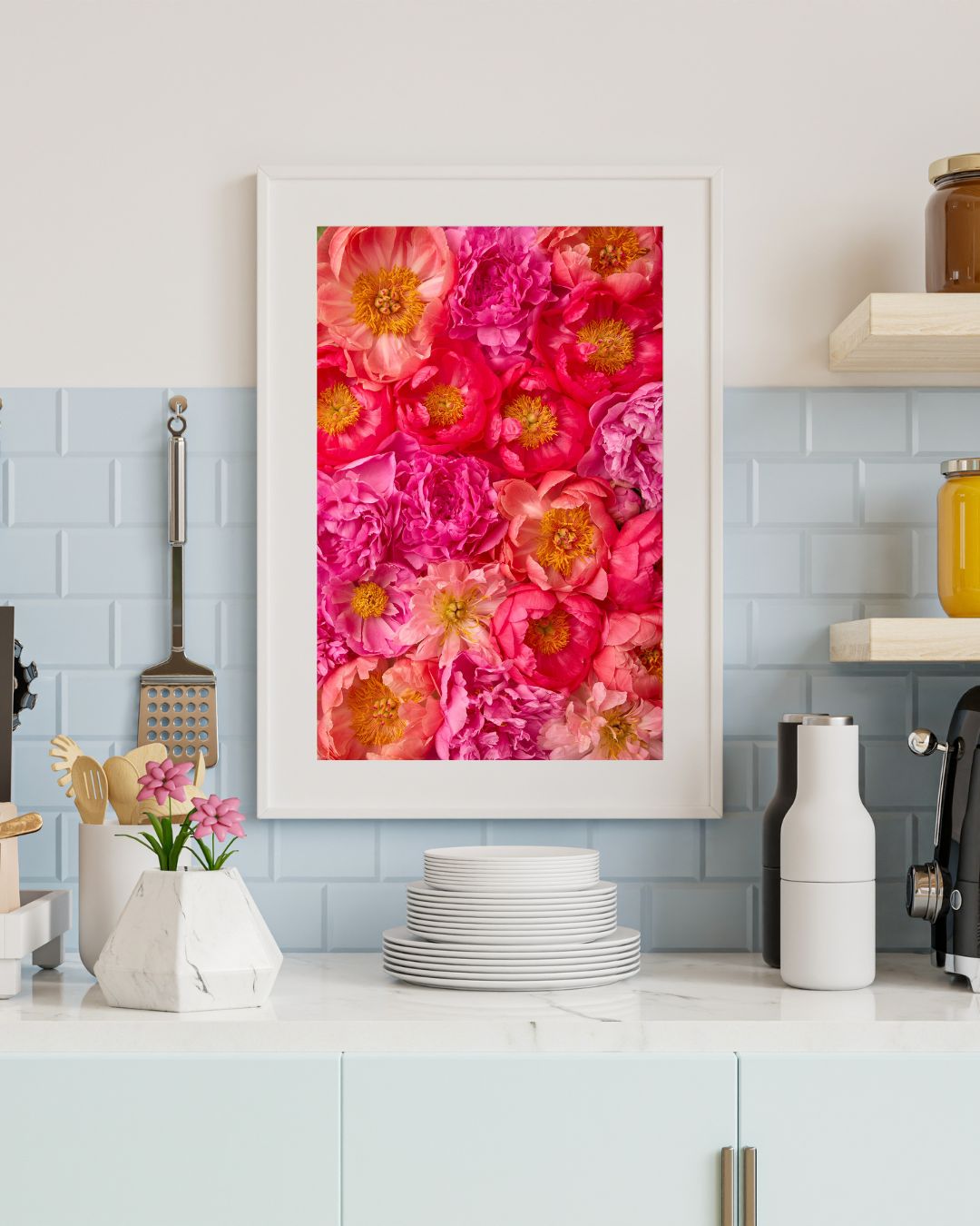 A kitchen counter with white tiles and light blue accents showcases the framed fine art paper print titled Peony Bed - Radiant Dreams, featuring pink and red flowers. Below, stacked plates and a marble utensil holder accompany jars and a coffee machine.