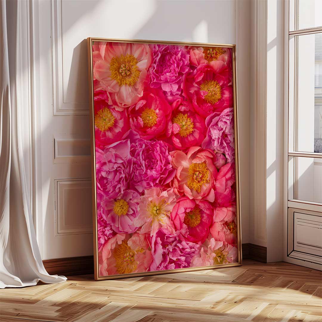 The Radiant Dreams large framed canvas, displaying vibrant pink and yellow flowers, leans against a white wall. Sunlight filters through a nearby window casting soft shadows on the wooden floor, while a white curtain partially drapes over it, adding peony perfection.