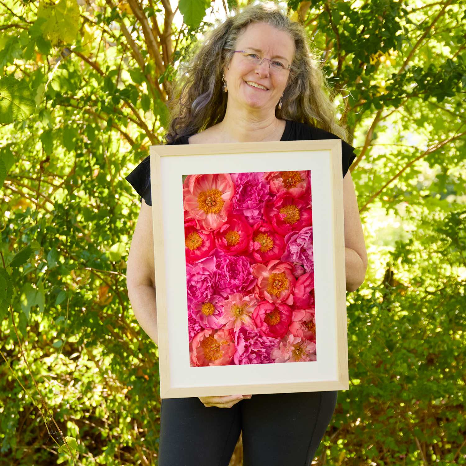 Christina Peters holding Radiant Dreams Peony Wall Art Print, a vertical close-up of coral, hot pink and magenta peonies packed together with golden centers showing, framed in natural wood with a white mat. She is standing outdoors in front of sunlit green trees and leafy shrubs.