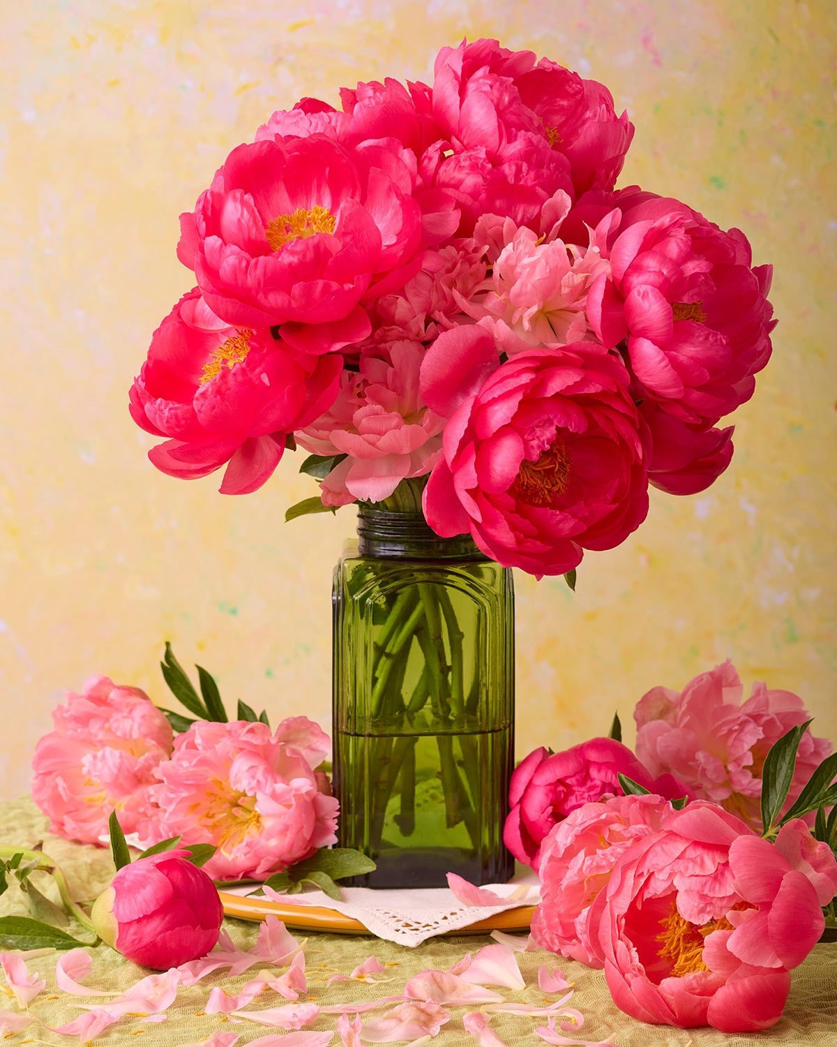 The Love Story Blooming features a vibrant coral-pink peony bouquet in a green glass vase, surrounded by scattered petals and blooms on a light green tablecloth, resembling exquisite canvas wall art against a soft yellow backdrop.