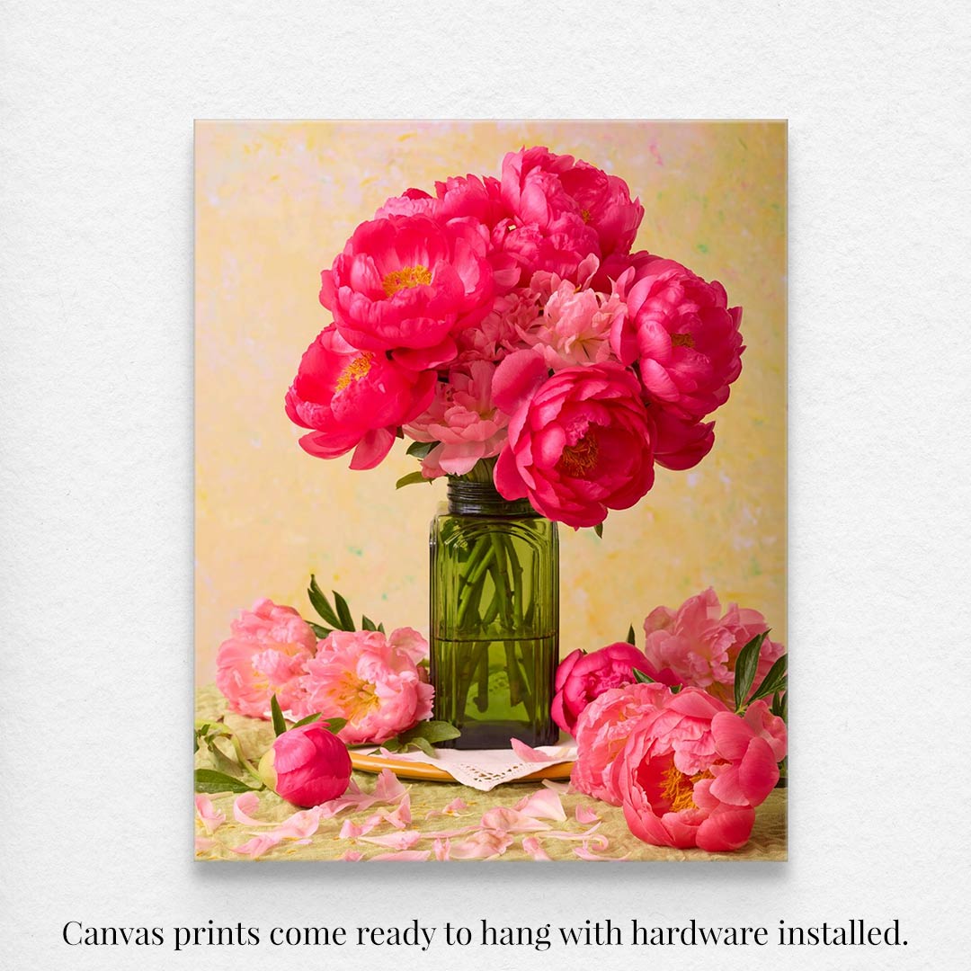 The Love Story Blooming canvas art showcases vibrant coral-pink peonies in a green glass jar with a soft yellow background and scattered fallen petals. It comes ready to hang with hardware installed.