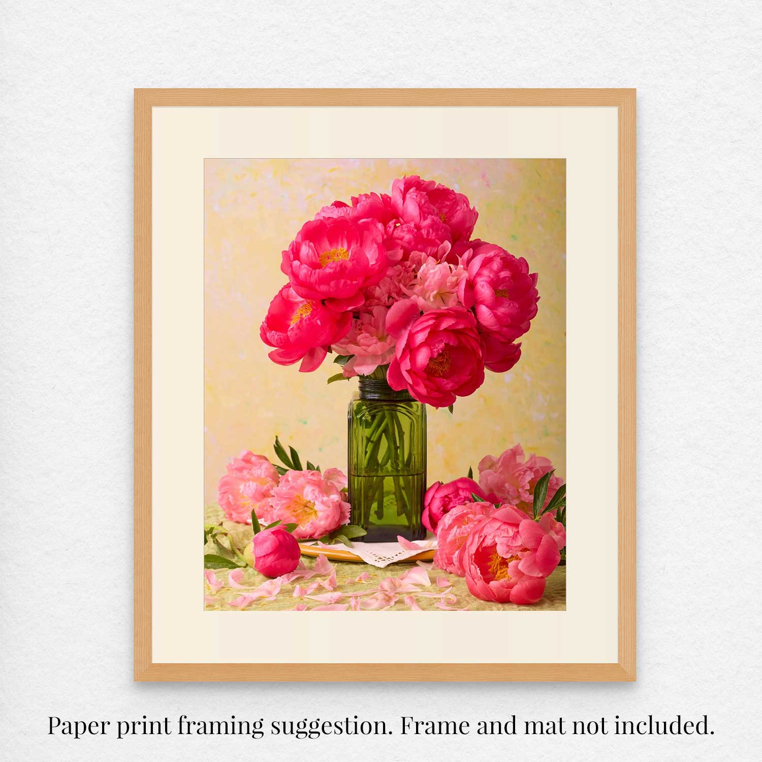 The Love Story Blooming print features coral-pink peonies in a green vase on a pastel backdrop, set in a simple wooden frame with a cream mat. Text below says: Paper print framing suggestion. Frame and mat not included.