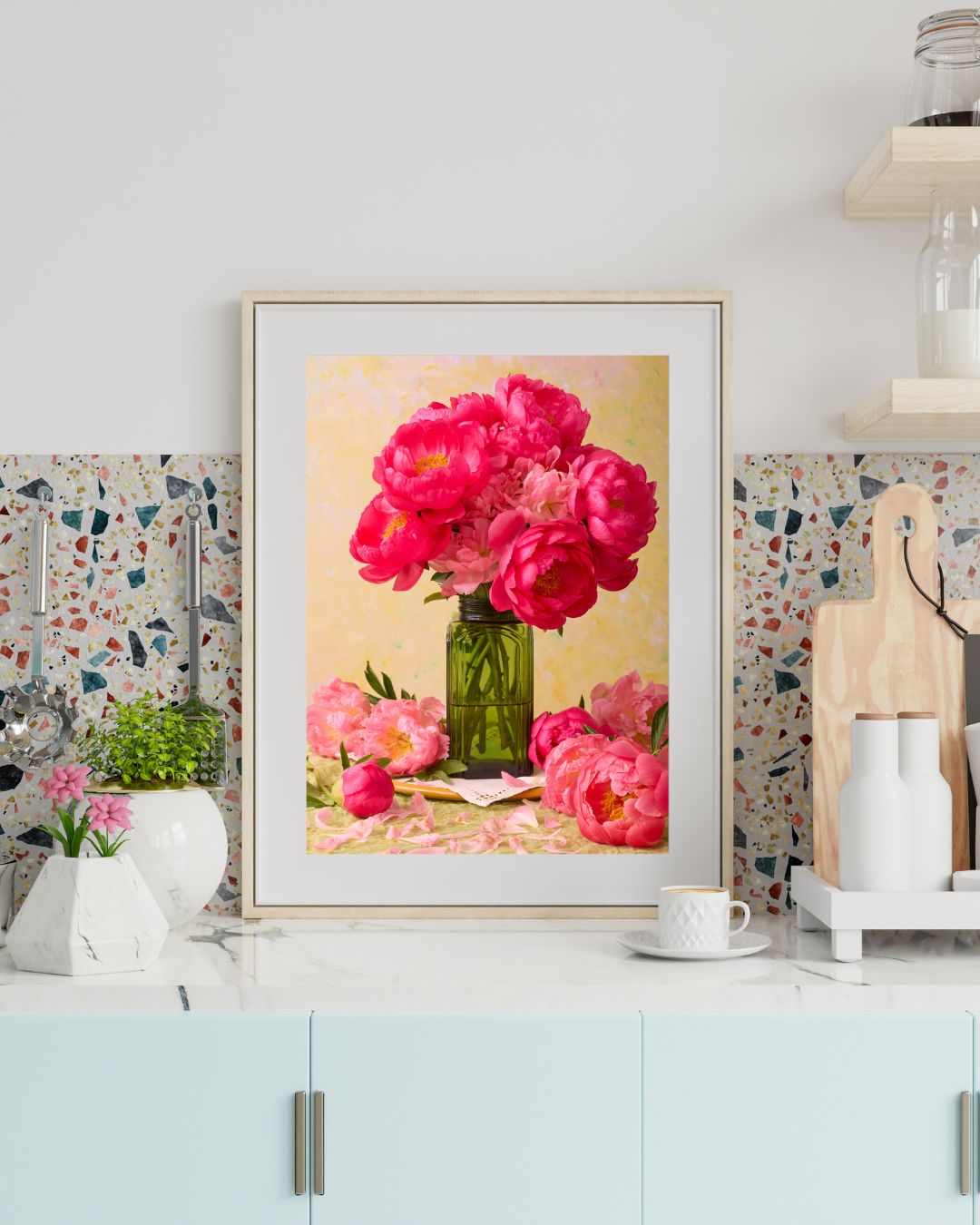 The Love Story Blooming framed, featuring vibrant coral-pink peonies in a green vase, adorns the kitchen counter. It sits alongside a potted plant, coffee cup, and cutting boards against the stylish terrazzo backsplash.