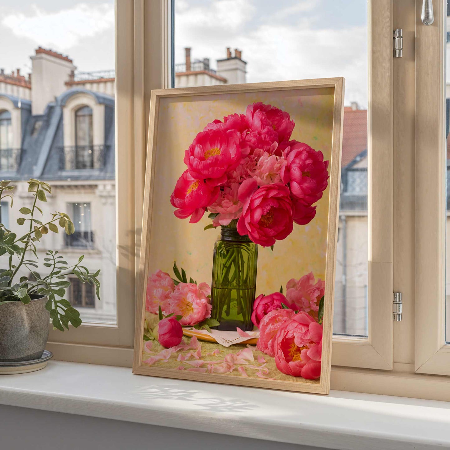 The framed Love Story Blooming canvas of coral-pink peonies in a green vase adorns the windowsill, with charming rooftops and chimneys adding a Parisian flair. A nearby potted plant enhances the serene scene.