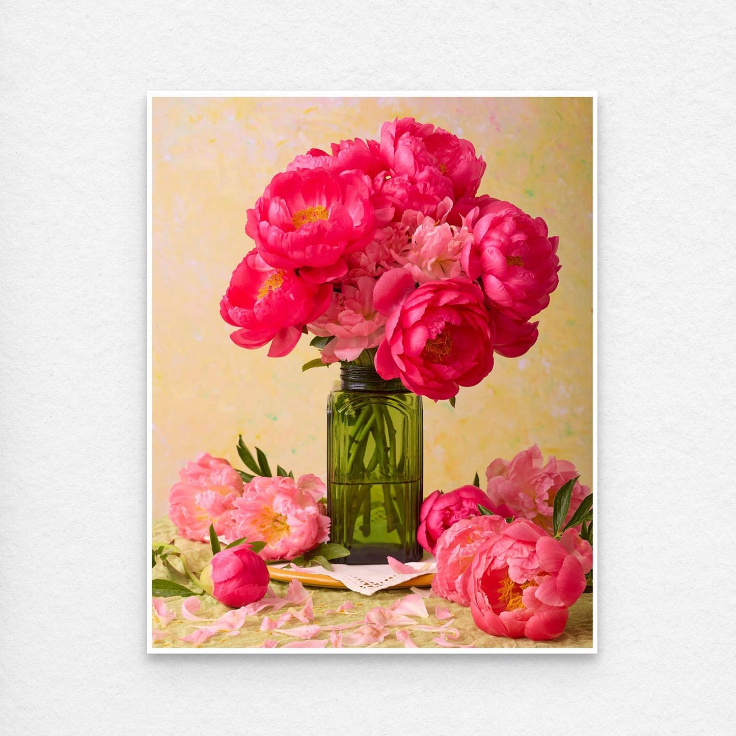 The Love Story Blooming features coral-pink peonies in a green vase on the table, surrounded by scattered petals. A soft, textured yellow background adds warmth to the scene, ideal for fine art paper prints or canvas wall art. This print is showing a thin white border, standard with all paper prints.