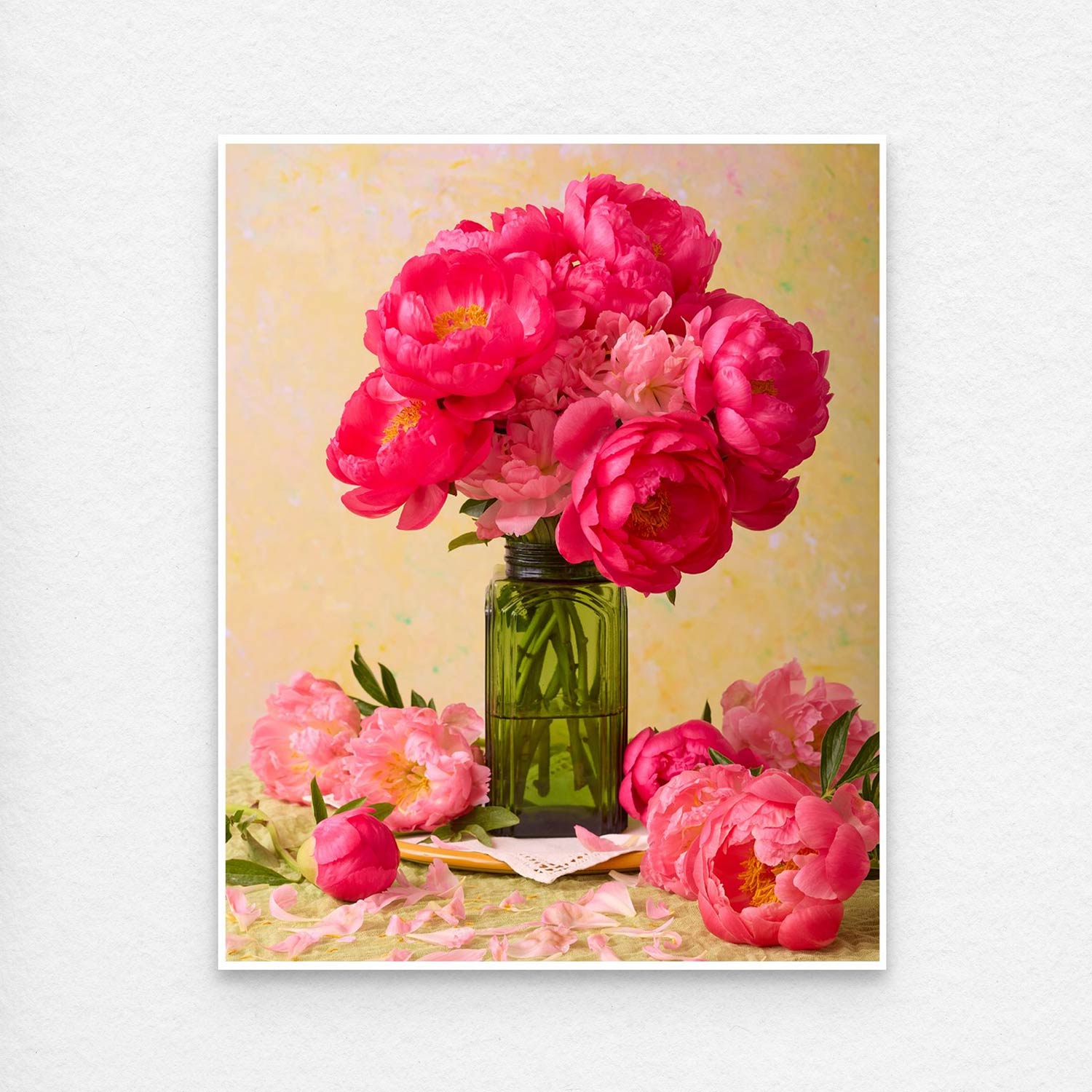 The Love Story Blooming features coral-pink peonies in a green vase on the table, surrounded by scattered petals. A soft, textured yellow background adds warmth to the scene, ideal for fine art paper prints or canvas wall art. This print is showing a thin white border, standard with all paper prints.