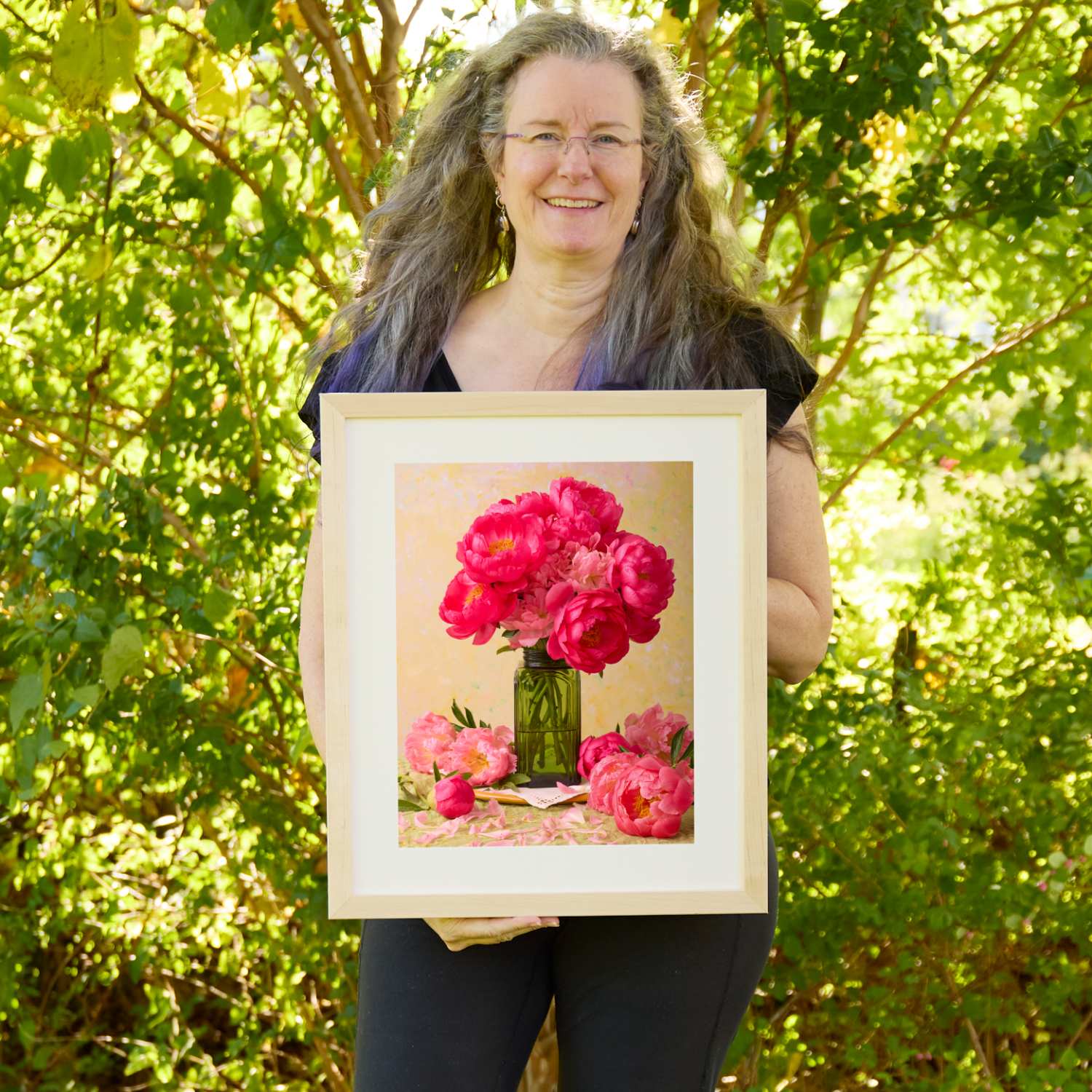 Christina Peters holding Love Story Blooming Peony Wall Art Print, a still life of vivid coral-pink peonies in a green glass jar, with loose blooms and petals around it against a soft golden backdrop, framed in natural wood with a white mat. She is standing outdoors in front of sunlit green trees and leafy shrubs.