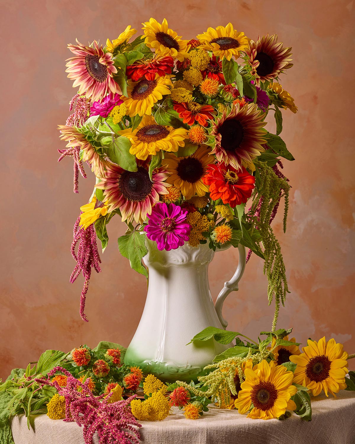 The Golden Fiesta features a vibrant mix of sunflowers, zinnias, and greenery cascading from a white ceramic vase. Yellow blooms spill elegantly onto the beige cloth below with a warm gradient background.