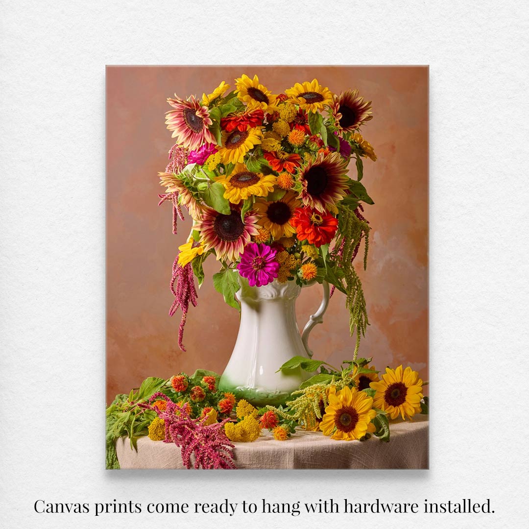 The Golden Fiesta canvas art features vibrant sunflowers and assorted flowers in a white ceramic pitcher against a soft brown background, with blossoms cascading elegantly over the tables edge. Canvas prints come ready to hang.