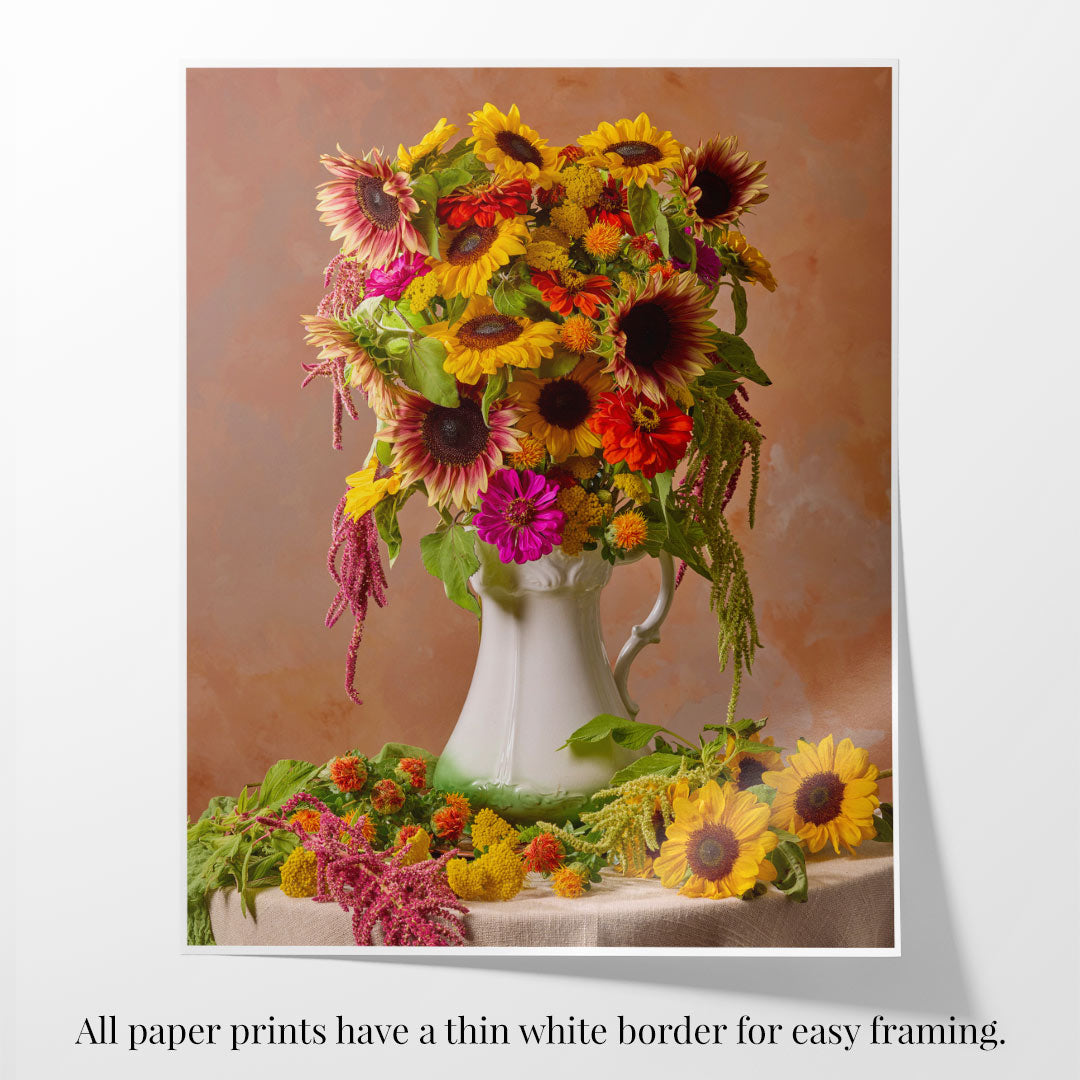 The Golden Fiesta features a vibrant bouquet of sunflowers, zinnias, and greenery in a white ceramic vase on a table, with additional flowers scattered at the base. Perfect for a fine art print, it comes with a thin white border for easy framing.