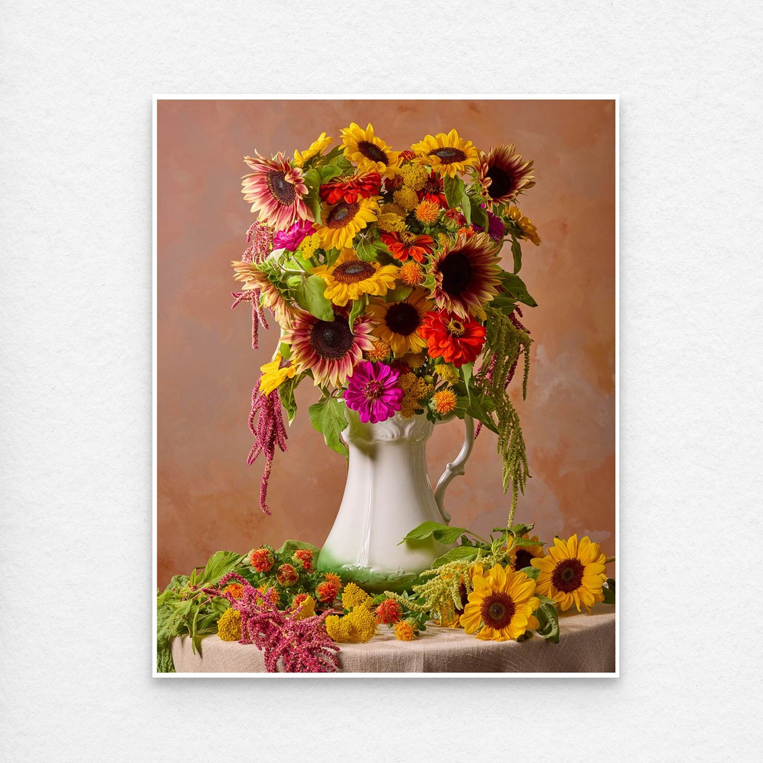 The Golden Fiesta bouquet, featuring sunflowers and vibrant blooms, is artfully arranged in a white pitcher against a warm brown backdrop. Additional flowers and greenery enhance the display. Paper print showing a white border. 