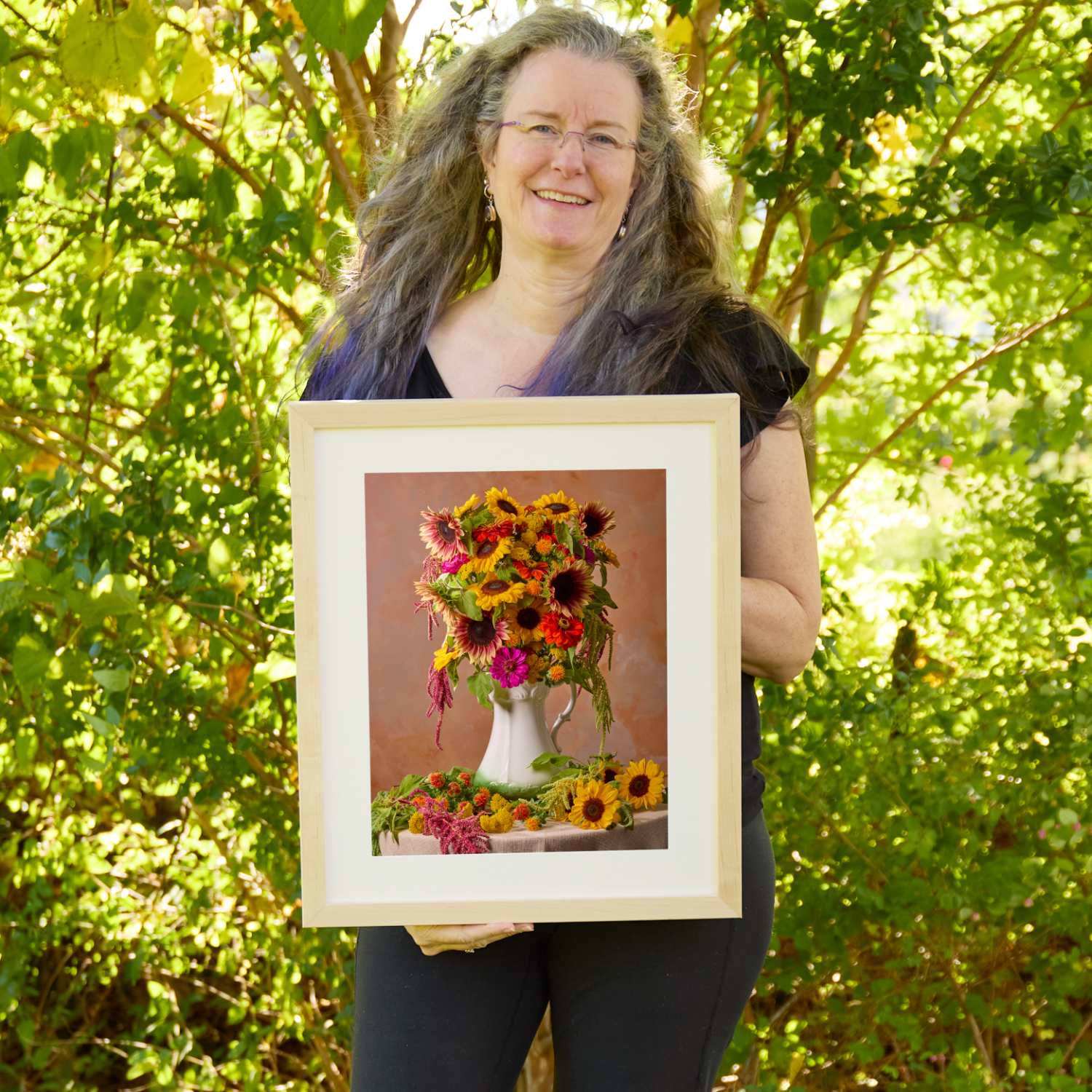 Christina Peters holding Golden Fiesta Sunflower Wall Art Print, a still life of golden and burgundy sunflowers, zinnias and trailing amaranth in a white pitcher, with more sunflowers on the table against a warm terracotta backdrop, framed in natural wood with a white mat. She is standing outdoors in front of sunlit green trees and leafy shrubs.