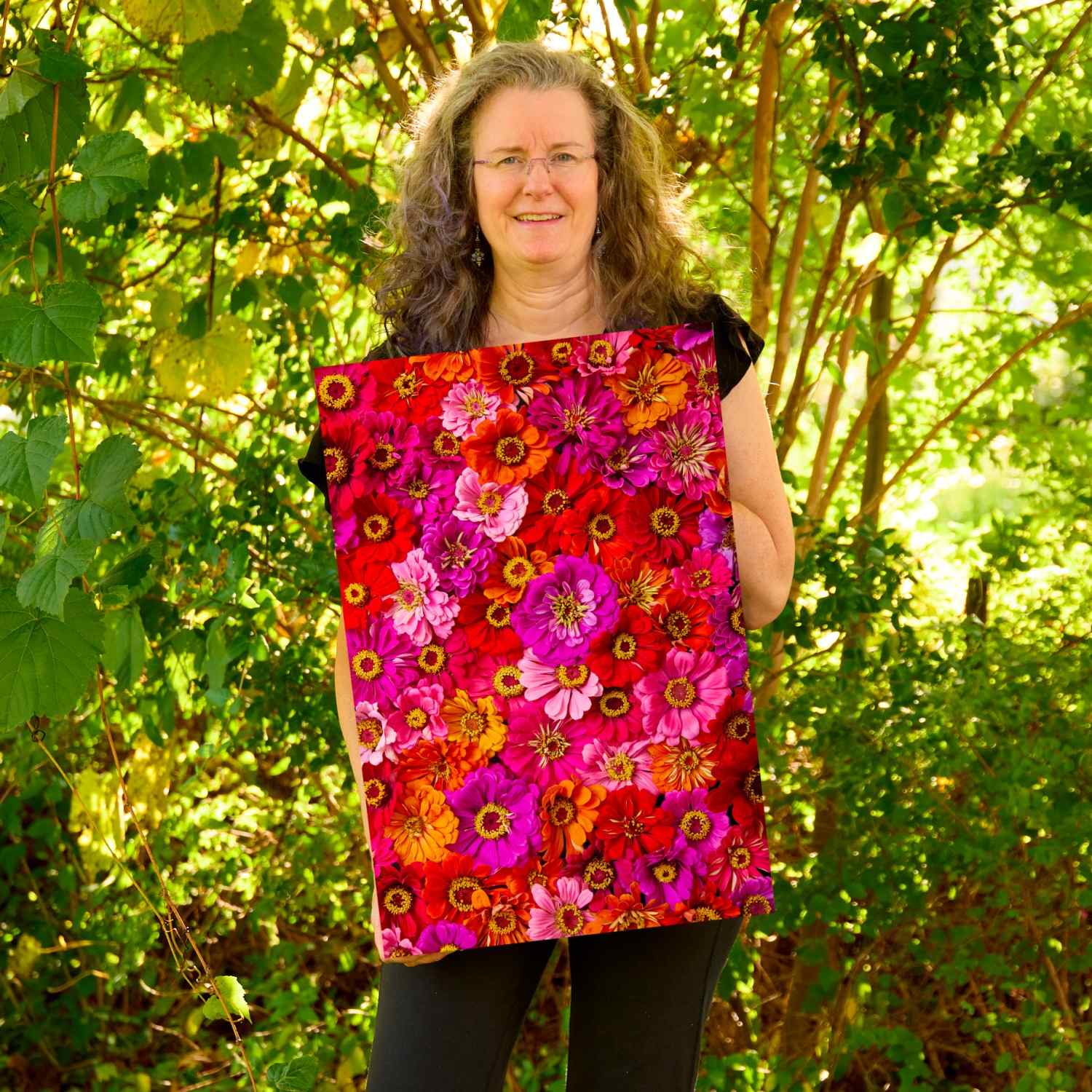 Christina Peters holding Zinnias Electric Radiance Wall Art Canvas, a vertical close-up of vivid red, magenta, pink and orange zinnias packed together. She is standing outdoors in front of sunlit green trees and leafy shrubs.