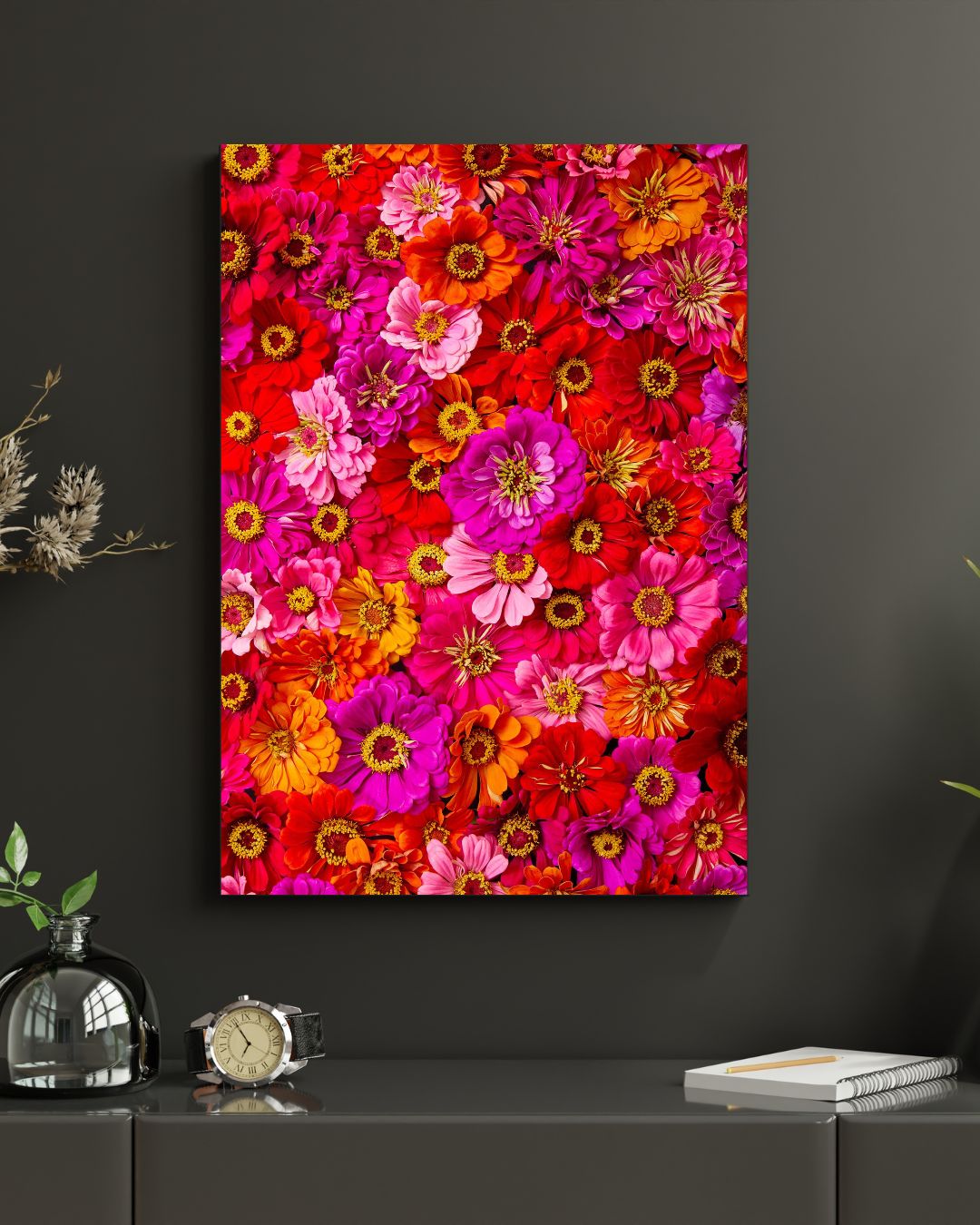 The Zinnias Electric Radiance canvas art displays pink, red, and orange zinnias on a dark wall. Below it sits a table with a modern glass vase holding small branches, alongside a wristwatch and stacked books.