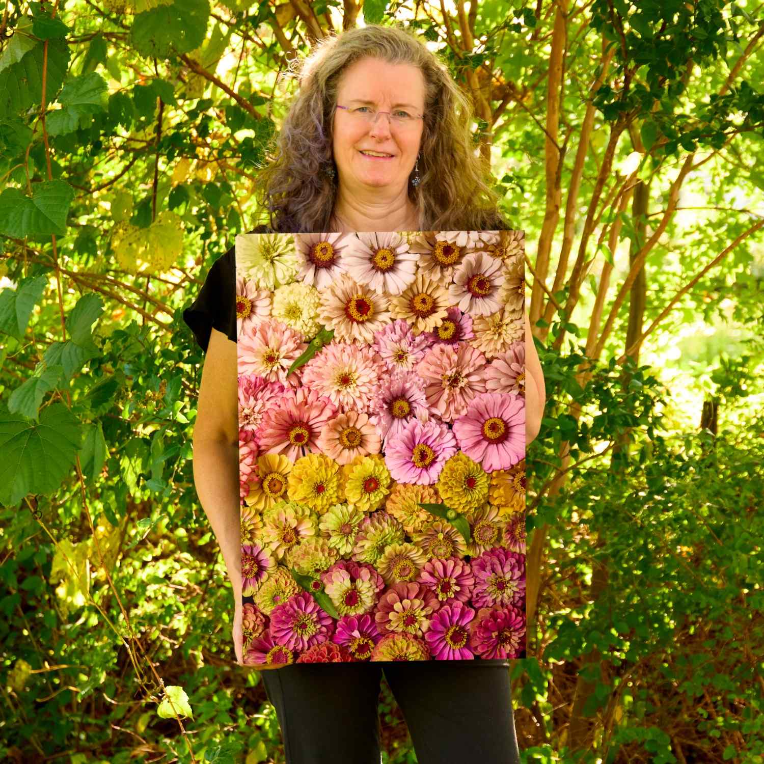 Christina Peters holding Zinnias Dawn To Dusk Wall Art Canvas, a vertical close-up of zinnias arranged in soft bands of pink, peach, gold and magenta. She is standing outdoors in front of sunlit green trees and leafy shrubs.