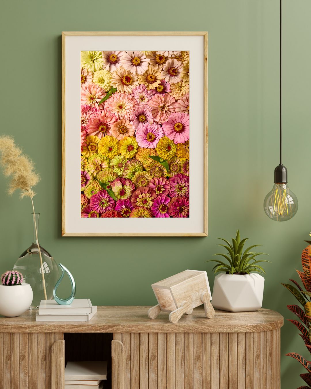 A minimalist room includes a wooden cabinet, a plant, book, glass ornament, and beige rocking horse. The green wall is adorned with the framed Zinnias Dawn To Dusk canvas art. A pendant light with an exposed bulb adds charm from the ceiling.