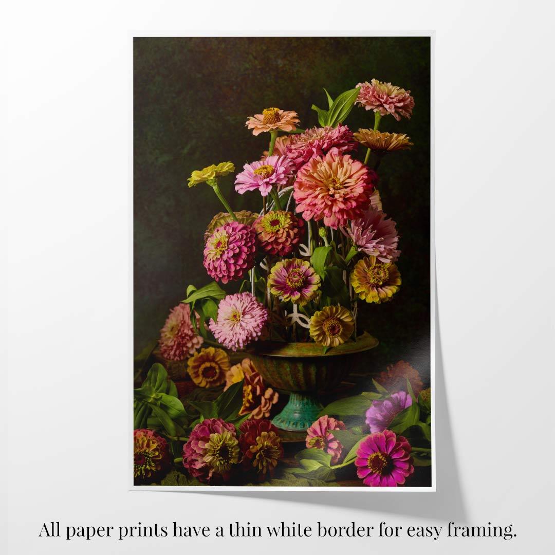 The Petals In Flight print features a vibrant bouquet of pink, yellow, and coral zinnias in a decorative vase against a dark backdrop, with text noting that all fine art paper prints include a thin white border for easy framing.