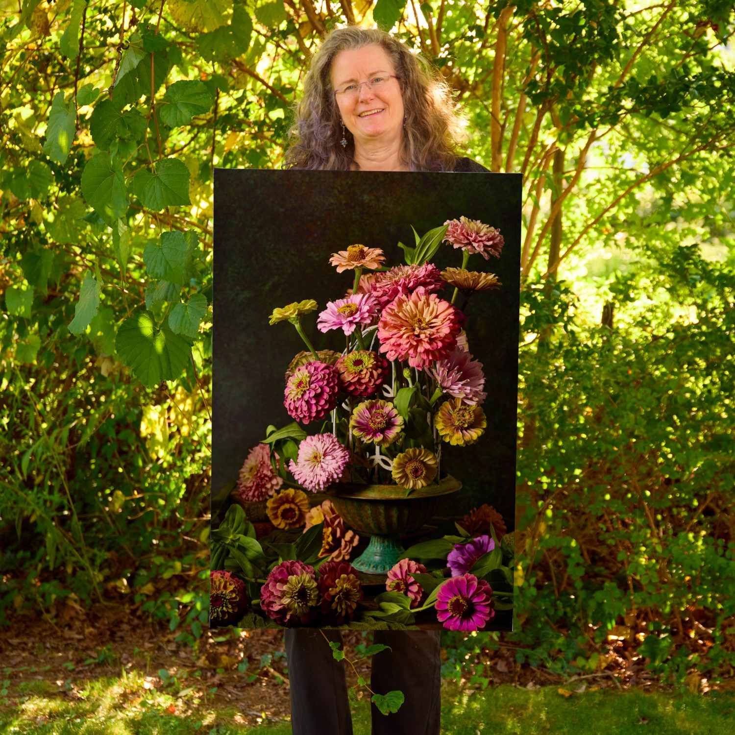 Christina Peters holding Petals In Flight Zinnia Fine Art Canvas, a dramatic still life of pink, coral and magenta zinnias in a footed teal bowl, with blooms spilling below against a deep black backdrop. She is standing outdoors in front of sunlit green trees and leafy shrubs.